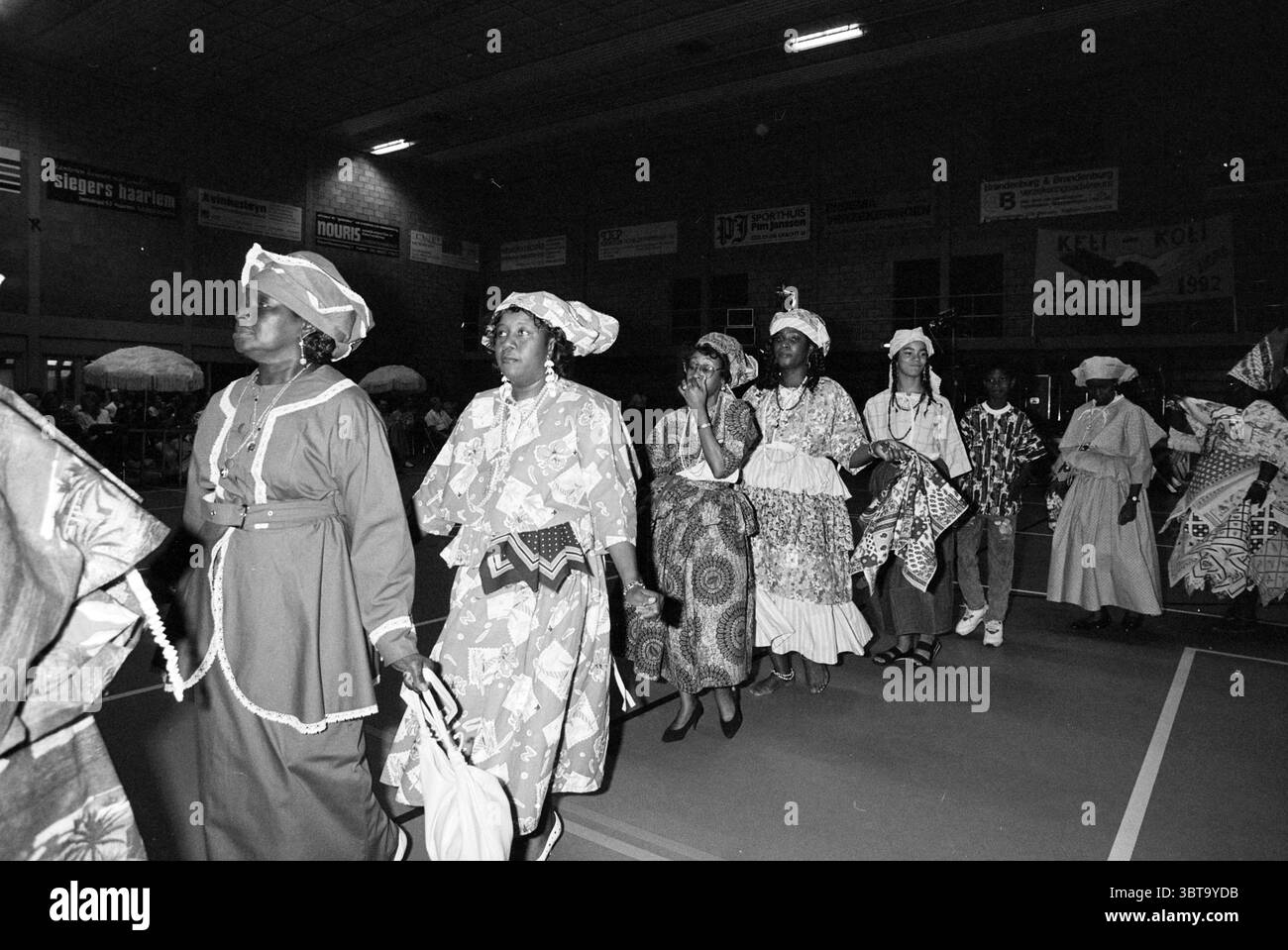 Tanzfestival im Beyneshal in Haarlem Haarlem Niederlande, Whizgle News, Dutch Desk, Niederlande, 1950 - 2000 am 26.06.1992. Die Abbildung zeigt diese Themen. Die Szene fängt ein lebendiges kulturelles Ereignis mit einer Prozession von Personen in traditioneller Kleidung ein. Jede Person trägt ein unverwechselbares Outfit mit komplizierten Mustern, das eine reiche Vielfalt an Stoff und Design zeigt. Ihre Kleidungsstücke haben oft helle Farben wie tiefes Rot, lebhaftes Blau und markantes weiß, die einen schönen Kontrast zu den gedämpften Tönen der Inneneinrichtung bilden. Die Beleuchtung ist weich und dennoch mit Stockfoto