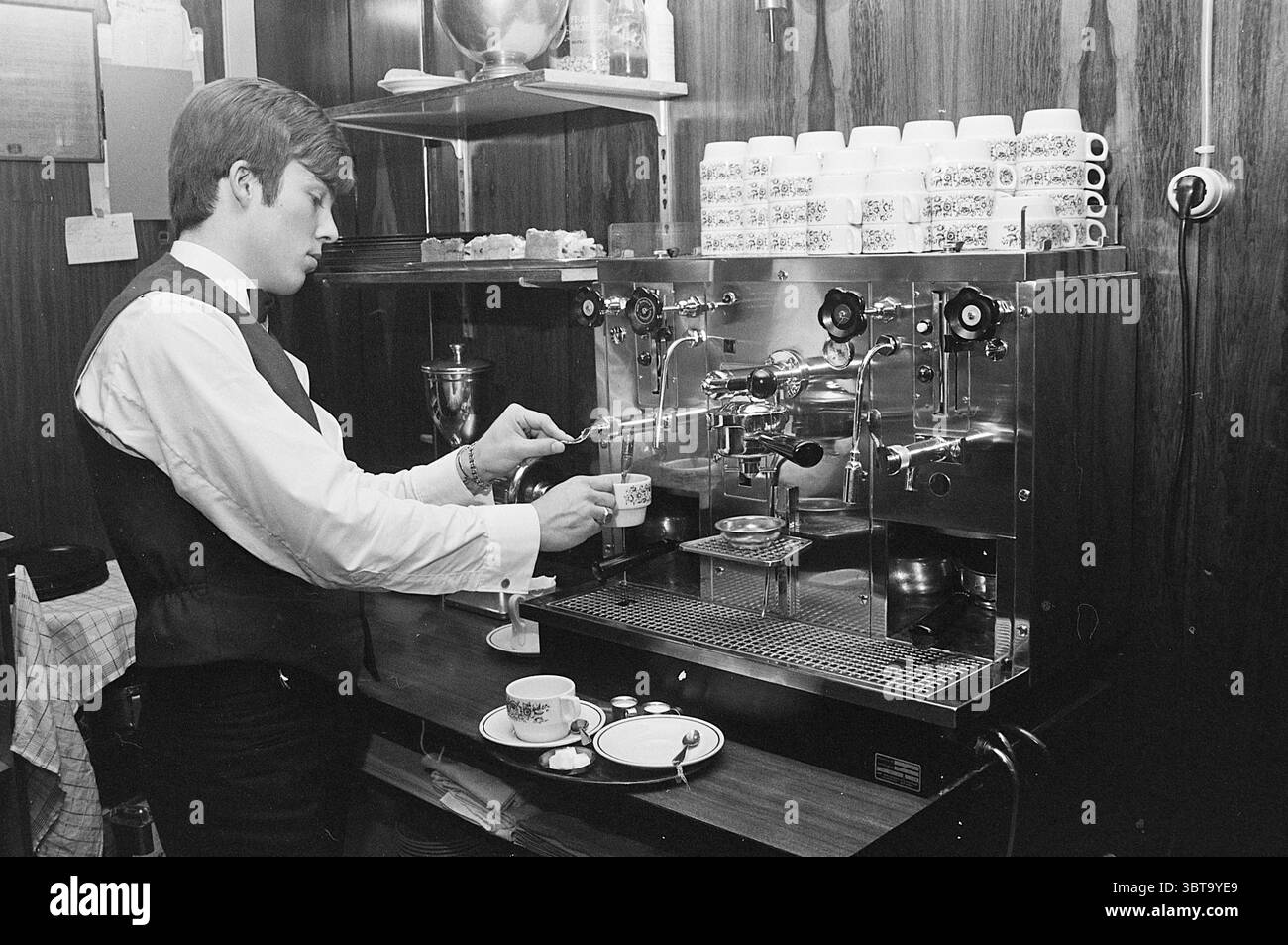 Report Coffee Report, Whizgle News, Dutch Desk, Niederlande, 1950 - 2000 am 23-10-1967. Diese Themen werden in der Abbildung angezeigt. In einem warmen, beleuchteten Raum steht ein junger Mann in formeller Kleidung, komplett mit Weste und weißem Hemd, fokussiert an einer Espressomaschine. Sein Haar ist klassisch gestylt und ergänzt das Vintage-Ambiente der Umgebung. Die Espressomaschine ist ein poliertes Metallstück mit verschiedenen Knöpfen und Hebeln, die das Licht reflektieren und ein kompliziertes Design präsentieren. Um den Barista herum ist die Wand mit Holztäfelungen verziert, die einen rustikalen Touch verleihen. Hinter ihm, eine Reihe von s Stockfoto