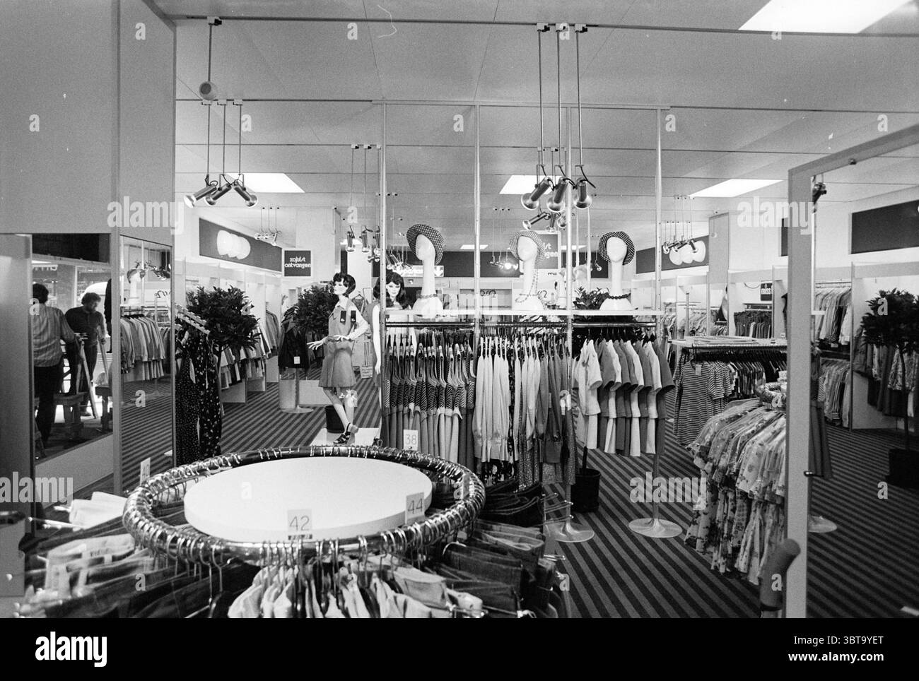 Innenraum von Voss in der Grote Houtstraat Innenraum Haarlem Grote Houtstraat Niederlande, Whizgle News, Dutch Desk, Niederlande, 1950 - 2000 am 06-05-1974. Dies sind die Elemente im Bild. Die Szene zeigt einen Verkaufsraum mit Kleiderständern und Displays. Im Mittelpunkt der Komposition steht ein kreisförmiger Kleiderständer, der im Vordergrund deutlich positioniert ist und von einer Vielzahl von Kleidungsstücken überflutet wird. Um dieses zentrale Element herum säumen zusätzliche Regale die Wände, in denen ordentlich angeordnete Hemden, Kleider und andere Kleidungsstücke ausgestellt sind. Die Atmosphäre vermittelt ein Gefühl der Ordnung, mit dem Stockfoto