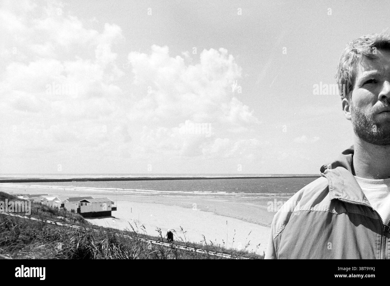 Herr Guustaaf Bakker Ijmuiden IJmuiden the Netherlands, Whizgle News, Dutch Desk, Niederlande, 1950 - 2000 am 02-07-1990. Das Bild enthält diese Themen. In dieser Szene steht ein Mann im Vordergrund, sein Ausdruck ist besinnlich. Er hat ein kurzes, zerzaustes Haar und trägt eine helle Jacke. Die Farbpalette ist gedämpft, dominiert von Grautönen, weiß und Anklängen gedämpfter Erdtöne. Hinter ihm erstreckt sich ein riesiger Strand, wo der Sand weich und leicht strukturiert erscheint. Die Küste biegt sich sanft in die Ferne und führt zu einem Horizont, der auf ein ruhiges Meer trifft. Stockfoto