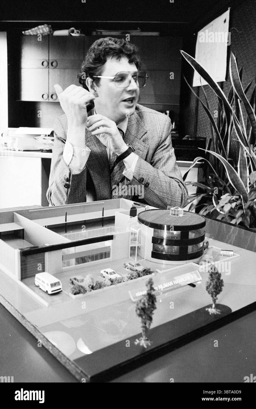 HR van Stein von der Firma Nijman + Model ( + Exterieur) Model Persons, Whizgle News, Dutch Desk, Niederlande, 1950 - 2000 am 05-03-1980. Das Bild enthält diese Themen. In einer gut beleuchteten Büroumgebung sitzt ein Mann nachdenklich an einem großen, modernen Schreibtisch. Er trägt einen hellen Anzug und eine runde Brille, die seinem Verhalten einen akademischen Eindruck verleihen. Sein Haar ist ordentlich gestylt, und er scheint in Gesprächen verwickelt zu sein und möglicherweise eine Idee oder ein Konzept zu präsentieren. Vor ihm befindet sich ein aufwendig gefertigtes Architekturmodell, das das Design eines Gebäudes mit klaren Linien und einer Mischung aus r zeigt Stockfoto