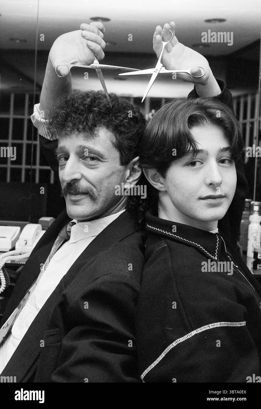 Friseure Don und Don Asser Haarlem Niederlande, Whizgle News, Dutch Desk, Niederlande, 1950 - 2000 am 16-11-1994. Das Bild enthält diese Themen. Die Szene fängt einen verspielten und humorvollen Moment zwischen zwei Personen ein, die Rücken an Rücken sitzen. Auf der einen Seite strahlt ein Mann mit einem hervorragenden Schnurrbart und einer lockigen Frisur Selbstvertrauen aus, gekleidet in einem maßgeschneiderten schwarzen Anzug, der seinem Verhalten einen Hauch von Professionalität verleiht. Sein Ausdruck ist warm und leicht schelmisch, was auf eine unbeschwerte Interaktion hindeutet. Auf der anderen Seite, eine jüngere Person mit glattem Haar, weich gestylt, Appeal Stockfoto