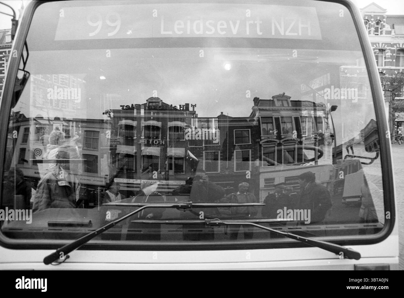 New NZH Bus GM, Whizgle News, Dutch Desk, Niederlande, 1950 - 2000 am 16-01-1992. Dies sind die Themen im Bild. Die Szene fängt eine lebendige urbane Umgebung ein, dominiert von einer großen, polierten Oberfläche – wahrscheinlich einem Busfenster –, das verschiedene Elemente um sie herum reflektiert. Im Vordergrund zeigt das Glas ein dynamisches Zusammenspiel von Reflexionen von Gebäuden und Menschen in der Umgebung. Der Bus selbst, der sich durch ein sauberes, weißes Äußere auszeichnet, steht vor dem Hintergrund historischer Architektur, mit detaillierten Fassaden und Fenstern, die auf ein reiches kulturelles Erbe hinweisen. Das können mehrere Personen Stockfoto