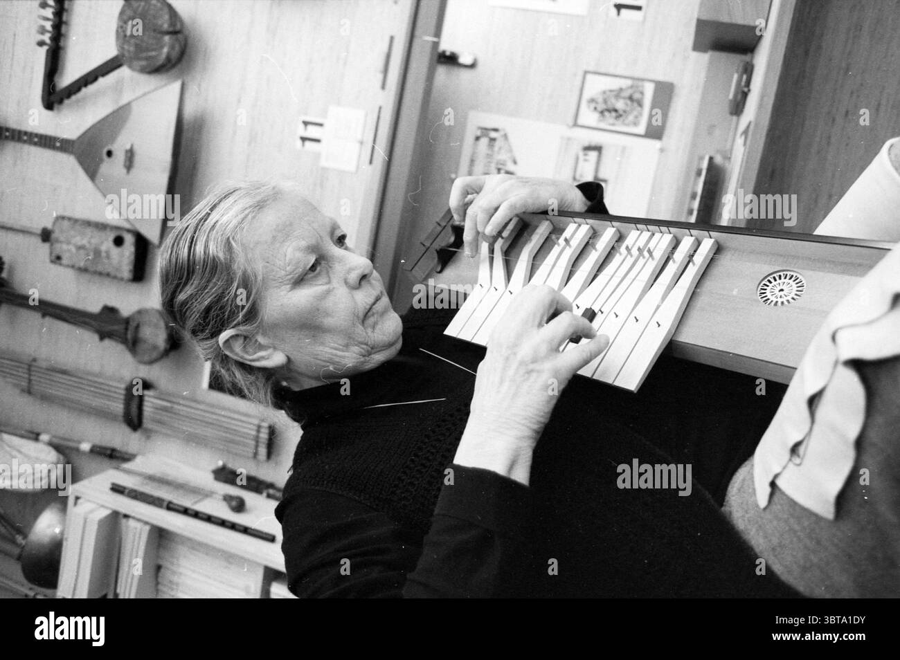 Frau van der Ree + alte Saiteninstrumente Bennebroek Bennebroek, Whizgle News, Dutch Desk, Niederlande, 1950 - 2000 am 13.02.1991. Das Bild enthält diese Themen. In einem warmen, sanft beleuchteten Innenraum spielt eine ältere Frau ein Holzinstrument, das einem Xylophon ähnelt. Sie hat silbergraue Haare ordentlich zurückgezogen, und ihr Ausdruck ist konzentriert und heiter, was auf eine tiefe Verbindung zur Musik hindeutet. In einem schwarzen Oberteil fügt sie sich harmonisch in die dezente, gemütliche Umgebung ein. Hinter ihr hängt eine Sammlung traditioneller Musikinstrumente an der Wand, einschließlich Stockfoto