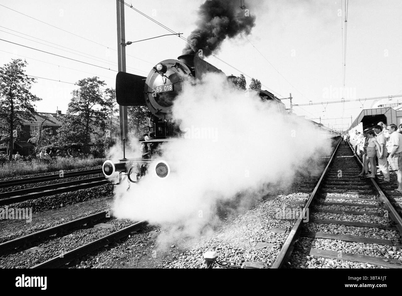 Ankunft Lokomotivbahnhof und Westergracht. Züge Haarlem Westergracht Niederlande, Whizgle News, Dutch Desk, Niederlande, 1950 - 2000 am 14-06-1986. Die Abbildung zeigt diese Themen. Die Szene zeigt eine klassische Dampflokomotive, die gut sichtbar auf Gleisen positioniert ist. Der Zug, der sich durch seine runde Front und Messingdetails auszeichnet, strahlt dicke, wellige Dampfwolken aus, die einen dramatischen und nostalgischen Effekt erzeugen. Der Kontrast zwischen dem tiefschwarzen Körper der Lokomotive und dem weißen Dampf erhöht die visuelle Intensität. Die Umgebung des Zuges bietet üppiges Grün mit Bäumen Stockfoto
