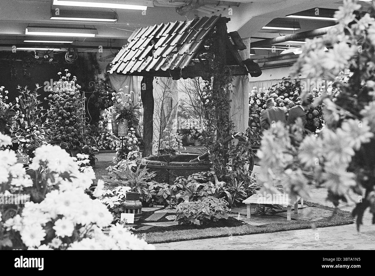 Blumenausstellung Aalsmeer Blumenausstellung, Whizgle News, Dutch Desk, Niederlande, 1950 - 2000 am 07-11-1968. Dies sind die Themen im Bild. Die Szene entfaltet sich in einer lebendigen Blumenausstellung, gefüllt mit einer Vielzahl blühender Pflanzen, die sorgfältig im gesamten Raum angeordnet sind. In der Mitte ist eine malerische Struktur, die einem traditionellen Brunnen ähnelt, aus altem Holz mit einem schrägen Dach mit dunklen Fliesen ausgestellt. Das Holz wirkt strukturiert und verwittert, was zu seinem rustikalen Charme beiträgt. Rund um den Brunnen sorgt eine Fülle von Blumen in verschiedenen Blütestadien für ein üppiges Atmo Stockfoto