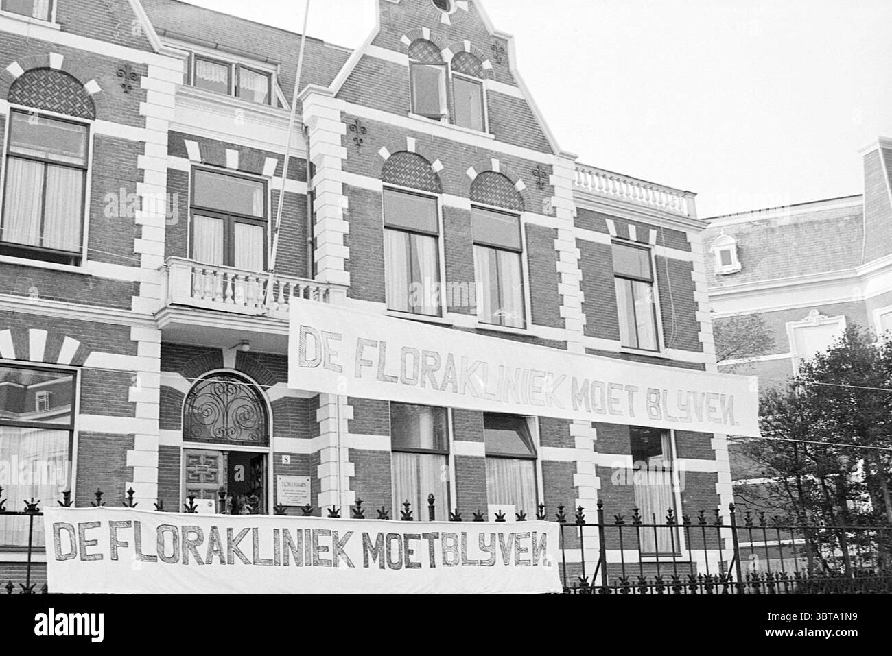 Erw. Flora Clinic Exterior, Whizgle News, Dutch Desk, Niederlande, 1950 - 2000 am 04-11-1973. Dies sind die Themen im Bild. Die Szene zeigt ein markantes Gebäude, das sich durch seinen viktorianischen Architekturstil auszeichnet. Die Fassade ist mit komplizierten Details wie dekorativen Zierleisten und großen, bogenförmigen Fenstern verziert. Die Struktur ist symmetrisch und weist einen zentralen Eingang auf, der von zusätzlichen Fenstern flankiert wird. Die Farbpalette ist überwiegend monochromatisch, mit Grau- und Weißtönen, was auf eine historische und etwas düstere Atmosphäre hindeutet. Banner werden gut sichtbar auf dem angezeigt Stockfoto
