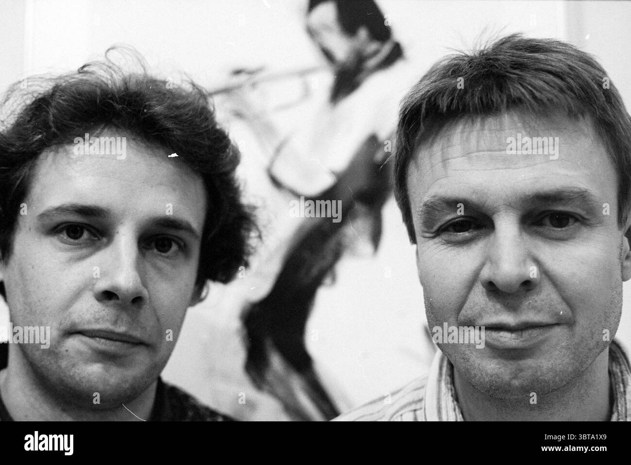 Eye-to-Eye Wispelberg + Ten Hoeve, Whizgle News, Dutch Desk, Niederlande, 1950 - 2000 am 15-12-1992. Dies sind die Elemente im Bild. Die Szene zeigt zwei Männer, die eng nebeneinander im Vordergrund stehen und beide direkt auf den Betrachter blicken. Der Mann auf der linken Seite hat lockiges Haar und einen nachdenklichen Ausdruck, während der Mann auf der rechten Seite glattes, helleres Haar und ein neutrales Verhalten hat. Ihre Gesichter sind sanft beleuchtet und bringen subtile Details in ihren Ausdrucksformen und Merkmalen hervor. Im Hintergrund ist eine stilisierte Darstellung eines Trompetenspielers zu sehen. Die Figur wird erfasst Stockfoto