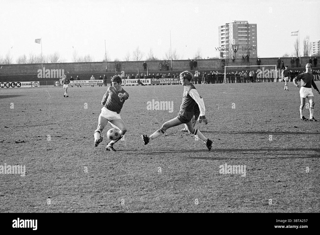 Fußballspiel FC Haarlem, Whizgle News, Dutch Desk, Niederlande, 1950–2000 im Jahr 1969. Das Bild enthält diese Themen. Die Szene fängt einen Moment auf einem grasbewachsenen Fußballfeld ein, auf dem zwei Spieler ein intensives Spiel erleben. Ein Spieler, der ein dunkles Trikot mit weißen Shorts trägt, ist bereit, den Ball zu kicken und sich mit Entschlossenheit nach vorne zu lehnen. Der zweite Spieler, der in helleren Farben gekleidet ist, hat gerade versucht, den Ball abzufangen. Sein Körper ist so positioniert, dass er Agilität und schnelle Reflexe suggeriert. Der Hintergrund zeigt eine Reihe von hohen Gebäuden, deren Fassaden einfach und Stockfoto