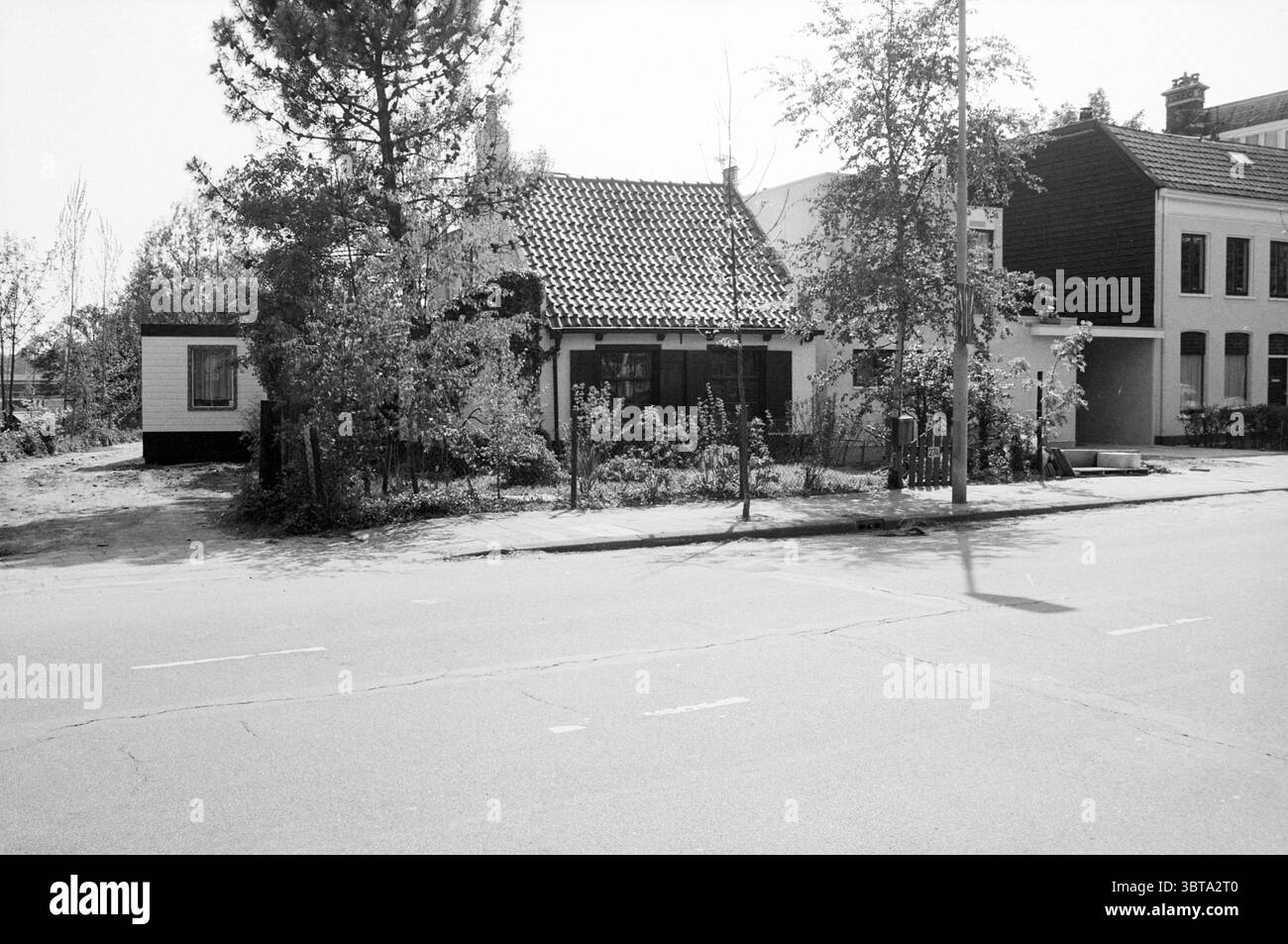 Notgebäude Zijlweg 274 Gebäude Haarlem Zijlweg, Whizgle News, Dutch Desk, Niederlande, 1950 - 2000 am 25-05-1987. Die Abbildung zeigt diese Themen. Die Szene zeigt ein malerisches einstöckiges Haus, das sich durch ein strukturiertes, geneigtes Ziegeldach und ein bescheidenes Äußeres in gedämpften Tönen auszeichnet. Um das Haus herum befinden sich verschiedene Grünflächen, darunter Büsche und kleine Bäume, die das gesamte Erscheinungsbild des Gebäudes mildern. Der Garten ist etwas wilde, was auf einen Mangel an neueren Instandhaltungsarbeiten hindeutet, wobei einige Pflanzen sichtbar auf den Wegen eindringen. Links ist eine kleinere Struktur. Stockfoto