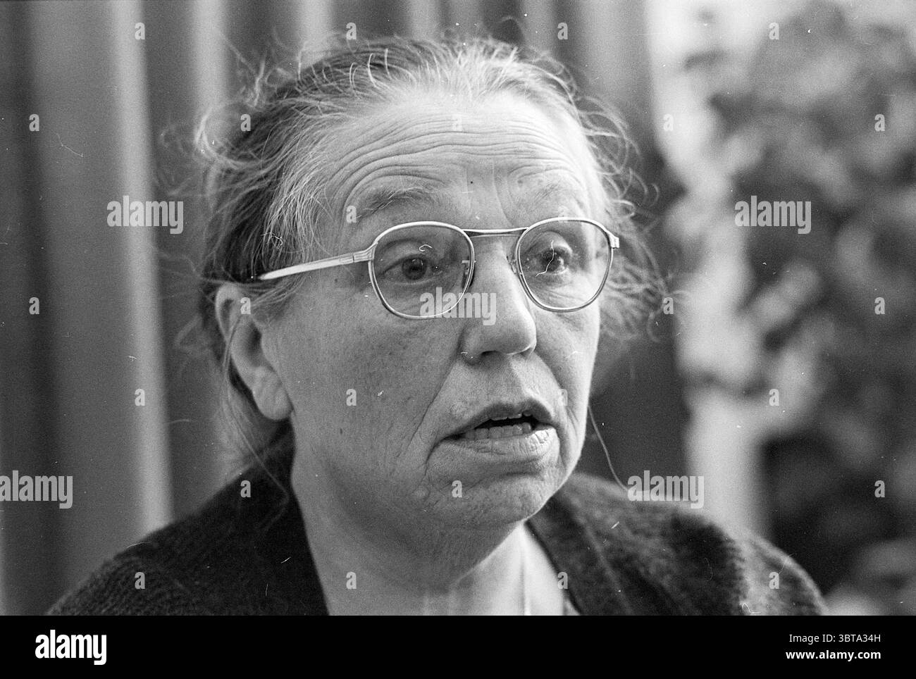 Frau de Haan ehemalige Haushälterin Menten Persons, Whizgle News, Dutch Desk, Niederlande, 1950 - 2000 am 26-11-1976. Das Bild enthält diese Themen. In der Szene wird eine ältere Frau mit einem ergreifenden Ausdruck dargestellt, der Besinnung oder Überraschung suggeriert. Sie hat ein dünnes, lang gezogenes Gesicht, das mit tief eingefassten Falten verziert ist und Geschichten von Erfahrung und Weisheit erzählt. Ihr graues Haar wird zurückgezogen und zeigt einen natürlich gealterten Teint und klar definierte Gesichtszüge. Sie trägt eine runde Brille, die leicht schräg auf der Nase sitzt und ihre ausdrucksstarken Augen verstärkt, die breit und aufmerksam wirken. Die Farben sind Stockfoto