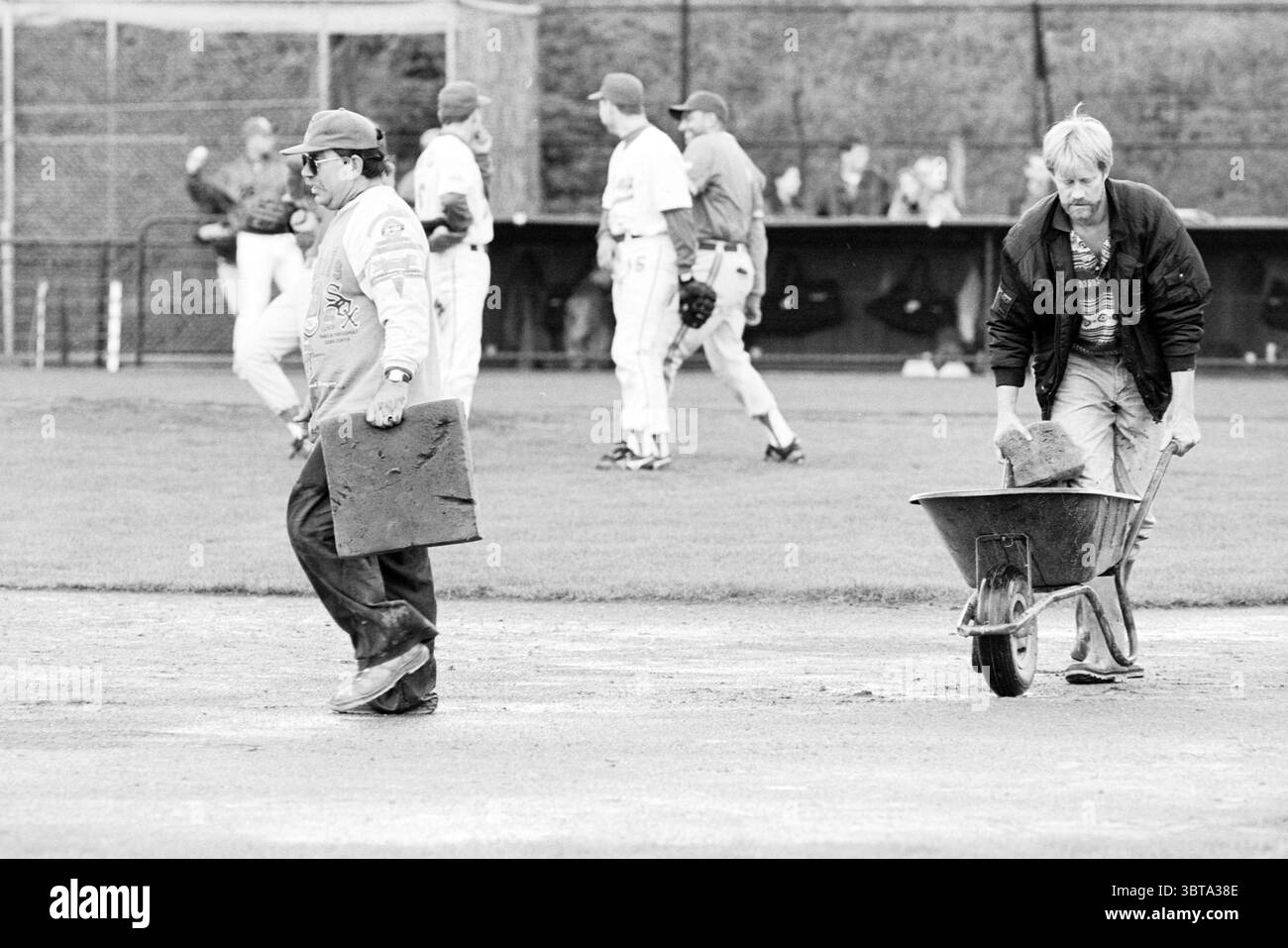 Rain Kinheim - HCAW, Whizgle News, Dutch Desk, Niederlande, 1950 - 2000 am 17.09.1994. Das Bild enthält diese Themen. In der Szene sind zwei Arbeiter im Vordergrund zu sehen, die jeweils unterschiedliche Aufgaben auf einem Sportplatz ausführen. Ein Arbeiter auf der linken Seite trägt eine dunkle Kappe und eine helle Jacke, die ein Quadrat aus Material trägt, das für Wartungsarbeiten verwendet wird, möglicherweise um einen beschädigten Bereich auf dem Feld abzudecken. Er geht zielgerichtet, fokussiert nach vorne und trägt zu einem Gefühl der Dringlichkeit seiner Bewegung bei. Der zweite Arbeiter, rechts positioniert, drückt ein Rad Stockfoto