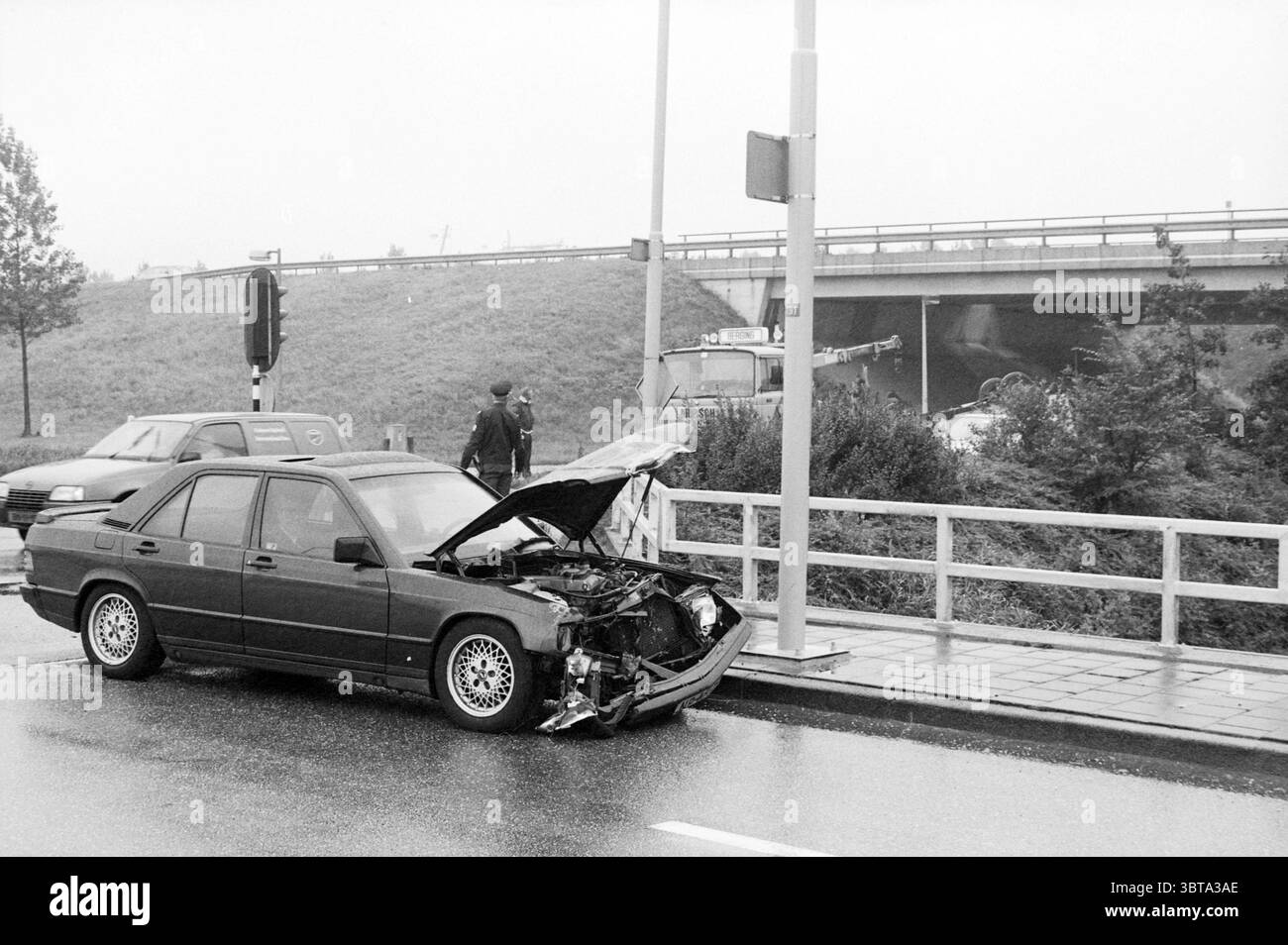 Autounfallwagen verkehrt herum, Whizgle News, Dutch Desk, Niederlande, 1950 - 2000 am 09-1988. Das Bild enthält diese Themen. Die Szene zeigt einen düsteren, regnerischen Tag auf einer Stadtstraße. Ein beschädigtes schwarzes Auto mit geöffneter Motorhaube steht nahe der Straßenkante und zeigt die verstümmelte Front, was auf eine kürzlich erfolgte Kollision hindeutet. Die Oberfläche des Fahrzeugs ist glatt vom Regen und reflektiert Tropfen und gedämpfte Farben. Auf der Straße sind einige Personen anwesend: Eine in dunkler Kleidung gekleidete untersucht das Auto mit einer gewissen Sorge, während eine andere Figur, möglicherweise ein Umstehender, in der Nähe steht, o Stockfoto