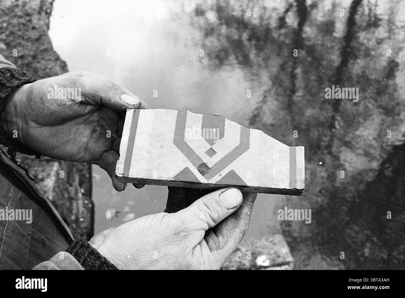 Alte historische Teichteiche, Whizgle News, Dutch Desk, Niederlande, 1950 - 2000 am 20-11-1969. Dies sind die Themen im Bild. Die Szene zeigt eine Nahaufnahme eines Handpaares, das ein kleines, rechteckiges Objekt, möglicherweise eine Fliese oder ein Stück Keramik, vor einem reflektierenden, wässrigen Hintergrund darstellt. Die Hände erscheinen gealtert und zeigen Anzeichen von Abnutzung und die Hautstruktur, die auf ein Leben mit manueller Arbeit hindeutet. Das Objekt selbst zeichnet sich durch ein markantes Design in gedämpften Farben aus – eine Mischung aus Erdtönen wie Rot-, Braun- und Cremetönen. Das geometrische Muster ist fett und etwas symmetrisch, was das Auge mit einem gewissen Augenmaß zeichnet Stockfoto