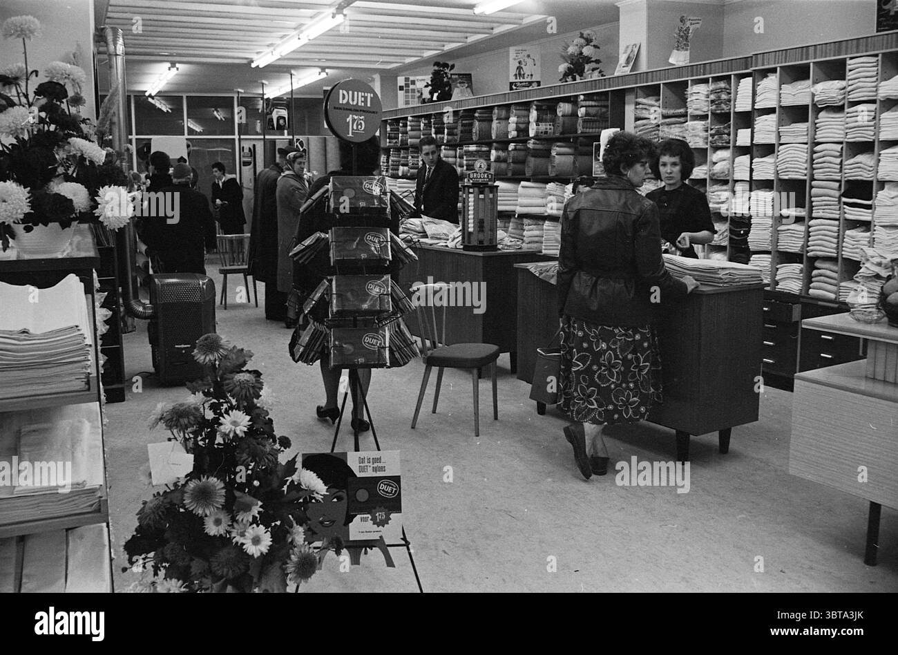 De Kroon kleine Houtstraat Interior Haarlem kleine Houtstraat Niederlande, Whizgle News, Dutch Desk, Niederlande, 1950 - 2000 am 18-11-1961. Das Bild enthält diese Themen. Die Szene fängt ein geschäftiges Einzelhandelsumfeld ein, wahrscheinlich ein Stoff- oder Kaufhaus, das mit verschiedenen Textilien gefüllt ist. Die Innenausstattung ist mit raumhohen Regalen ausgestattet, die mit fein gefalteten Stoffen bestückt sind und eine Reihe von Farben und Mustern, von einfarbigen Tönen bis hin zu Blumenmustern, präsentieren. Die Regale schaffen eine optisch ansprechende Kulisse mit einem organisierten, aber dennoch lebendigen Display. Im Vordergrund links befindet sich eine Anzeige des Pflanzenständers Stockfoto