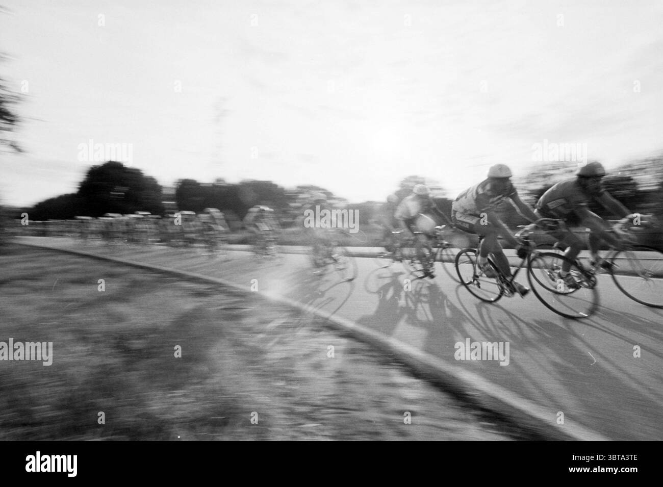 Radtour The Champion, Whizgle News, Dutch Desk, Niederlande, 1950 - 2000 am 16-08-1992. Das Bild enthält diese Themen. Die Szene erfasst einen dynamischen Moment eines Radrennereignisses, bei dem mehrere Radfahrer in Bewegung sind und ein Gefühl von Geschwindigkeit erzeugen. Die Fahrer tragen bunte Trikots, eine Mischung aus leuchtenden Rot-, Blau-, Grün- und Gelbtönen, die einen starken Kontrast zur monochromatischen Kulisse bilden. Der Hintergrund ist leicht verschwommen, was ein Gefühl schneller Bewegung hervorruft, während die Radfahrer eine Kurve umrunden. Der Boden scheint gepflastert zu sein, die Ränder sind von üppigem Grün geprägt, darunter dunkle, Stockfoto