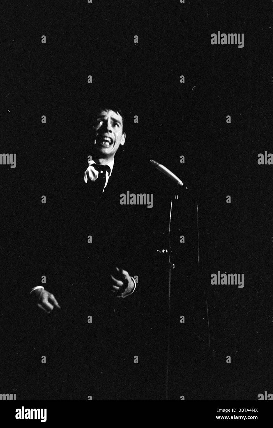 Aufführung von Jacques Brel, Whizgle News, Dutch Desk, Niederlande, 1950 - 2000 im Jahr 1963. Diese Themen werden in der Abbildung angezeigt. Die Szene zeichnet sich durch einen hohen Kontrast zwischen Licht und Schatten aus, wodurch eine dramatische Atmosphäre entsteht. Ein Künstler steht in der Mitte, beleuchtet von einem Scheinwerfer, der einen fokussierten Strahl auf seine Figur wirft, während die Umgebung in Dunkelheit verblasst. Die Person wirkt engagiert und animiert, vielleicht mitten im Satz, mit offenem Mund. Er trägt formelle Kleidung, die aus einem dunklen Anzug besteht, der sich in die umgebenden Schatten einfügt und die Intensität verstärkt Stockfoto