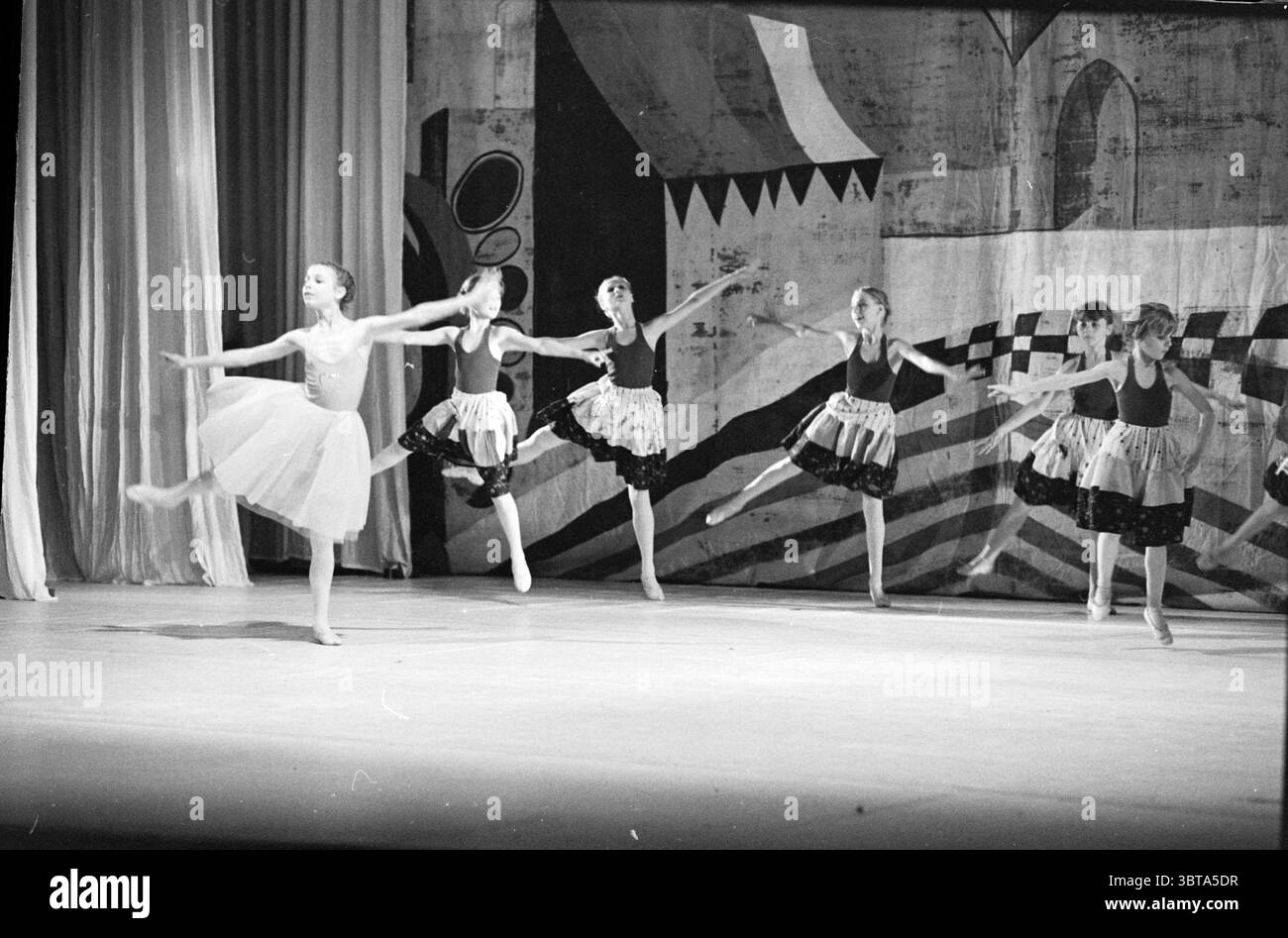 Ballettvorstellung Jugend, Whizgle News, Dutch Desk, Niederlande, 1950 - 2000 am 05-1981. Diese Themen sind in der Abbildung dargestellt. In einem von sanftem Licht getauchten Bühnenbild tritt eine Gruppe junger Tänzer auf, die ihre anmutigen Bewegungen und ihre Ausgeglichenheit zeigen. Der Fokus liegt auf einer zentralen Figur, die elegant auf einem Bein steht und Balance und Konzentration verkörpert. Diese Tänzerin trägt ein leichtes, fließendes Tutu, das im Kontrast zu den dunkleren Röcken der anderen Tänzerinnen steht, die sie umgeben und eine synchronisierte Formation beibehalten. Der Hintergrund zeigt ein abstraktes Wandbild mit geometrischen Formen und gedämpften, monochromatischen Tönen Stockfoto
