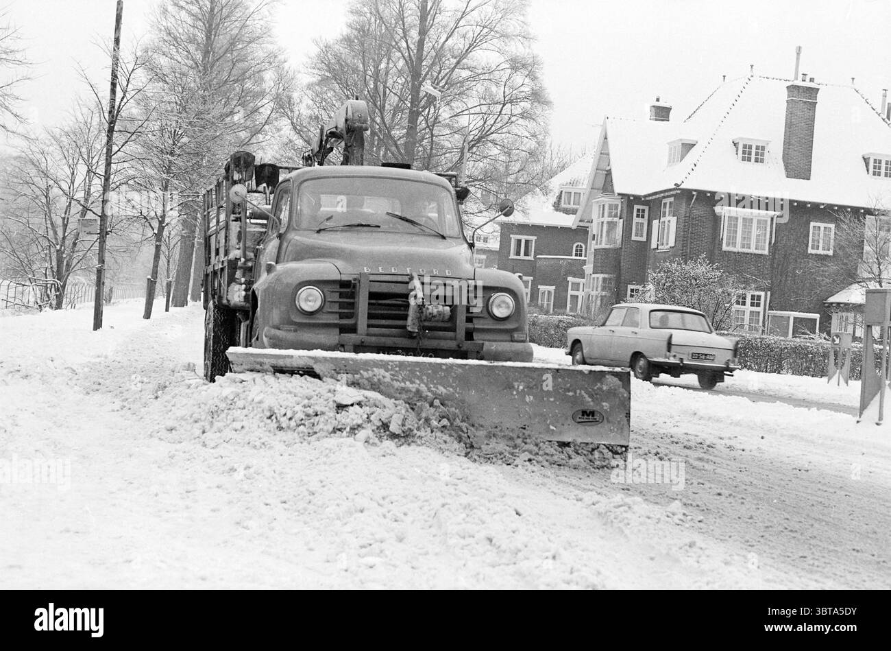 Schneepflug in Aktion in Haarlem., Whizgle News, Dutch Desk, Niederlande, 1950 - 2000. Diese Themen sind in der Abbildung dargestellt. Die Szene fängt eine Winterlandschaft mit frischem, pulverförmigem Schnee ein und schafft eine ruhige und dennoch krasse Atmosphäre. Ein Schneepflug nimmt den Vordergrund ein, dessen schweres, metallisches Blatt nach vorne geneigt ist und den Schnee beiseite drückt, um einen klareren Weg entlang der Straße zu schaffen. Der Lkw hat ein robustes Vintage-Design und seine Karosserie ist in einem gedämpften Ton lackiert, möglicherweise blau oder grün, was im Kontrast zur weißen Umgebung steht. Im Hintergrund stehen schneebedeckte Bäume hoch, ihre Äste er Stockfoto