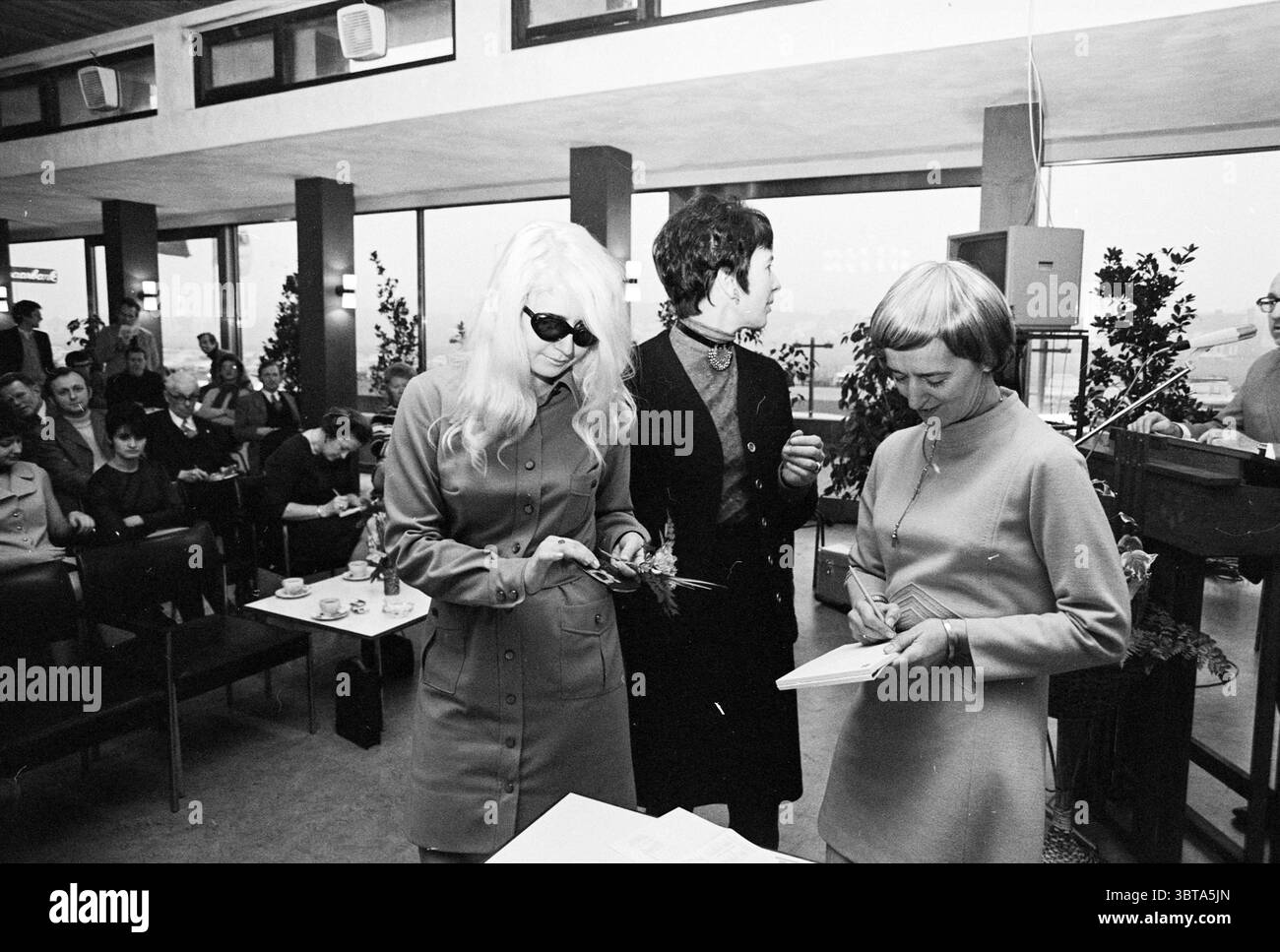 Drei Damen in einem Zimmer. Whizgle News, Dutch Desk, Niederlande, 1950 - 2000. Diese Themen werden in der Abbildung angezeigt. Die Szene fängt eine geschäftige Inneneinrichtung ein, die sich durch eine moderne Ästhetik aus der Mitte des Jahrhunderts auszeichnet. Der Raum verfügt über große Fenster, die natürliches Licht hereinlassen und den Raum mit einem weichen, diffusen Licht beleuchten. Topfpflanzen sind strategisch platziert und verleihen der ansonsten neutralen Umgebung einen Hauch von Grün und Wärme. Im Vordergrund stehen drei Frauen. Eine Frau mit langen, fließenden blonden Haaren, einer dunklen Sonnenbrille und einem stilvollen Mantel mit maßgeschneiderter Silhouette Stockfoto
