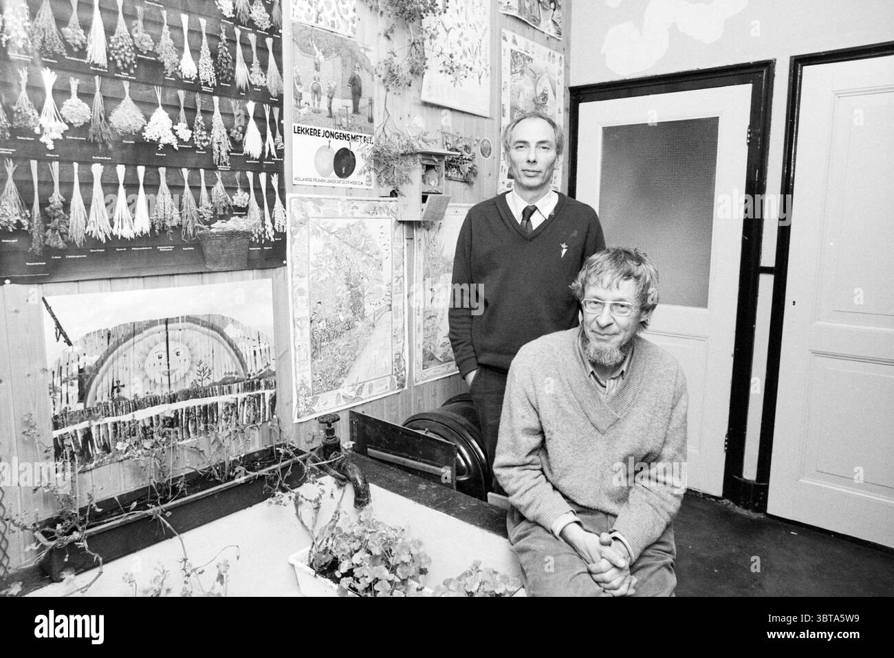 Herr Frits Darlang und Herr Ton Engelman vom Heimanshof Hoofddorp Wieger Bruinlaan, Niederlande., Whizgle News, Dutch Desk, Niederlande, 1950 - 2000. Diese Themen werden in der Abbildung angezeigt. In einem warm beleuchteten Innenraum stehen und sitzen zwei Männer neben einer Wand, die mit einer eklektischen Sammlung dekorativer Elemente und Kunstwerke geschmückt ist. Die Kulisse besteht aus einer Reihe lebendiger, detaillierter Karten und Illustrationen, die die Szene mit einem reichhaltigen visuellen Wandteppich versehen. Ein Mann steht mit ruhigem Verhalten, trägt einen dunklen Pullover über einem Hemd mit Kragen, seine Haare ordentlich gestylt. Er blickt mit einem leichten Lächeln auf den Zuschauer zu. Stockfoto