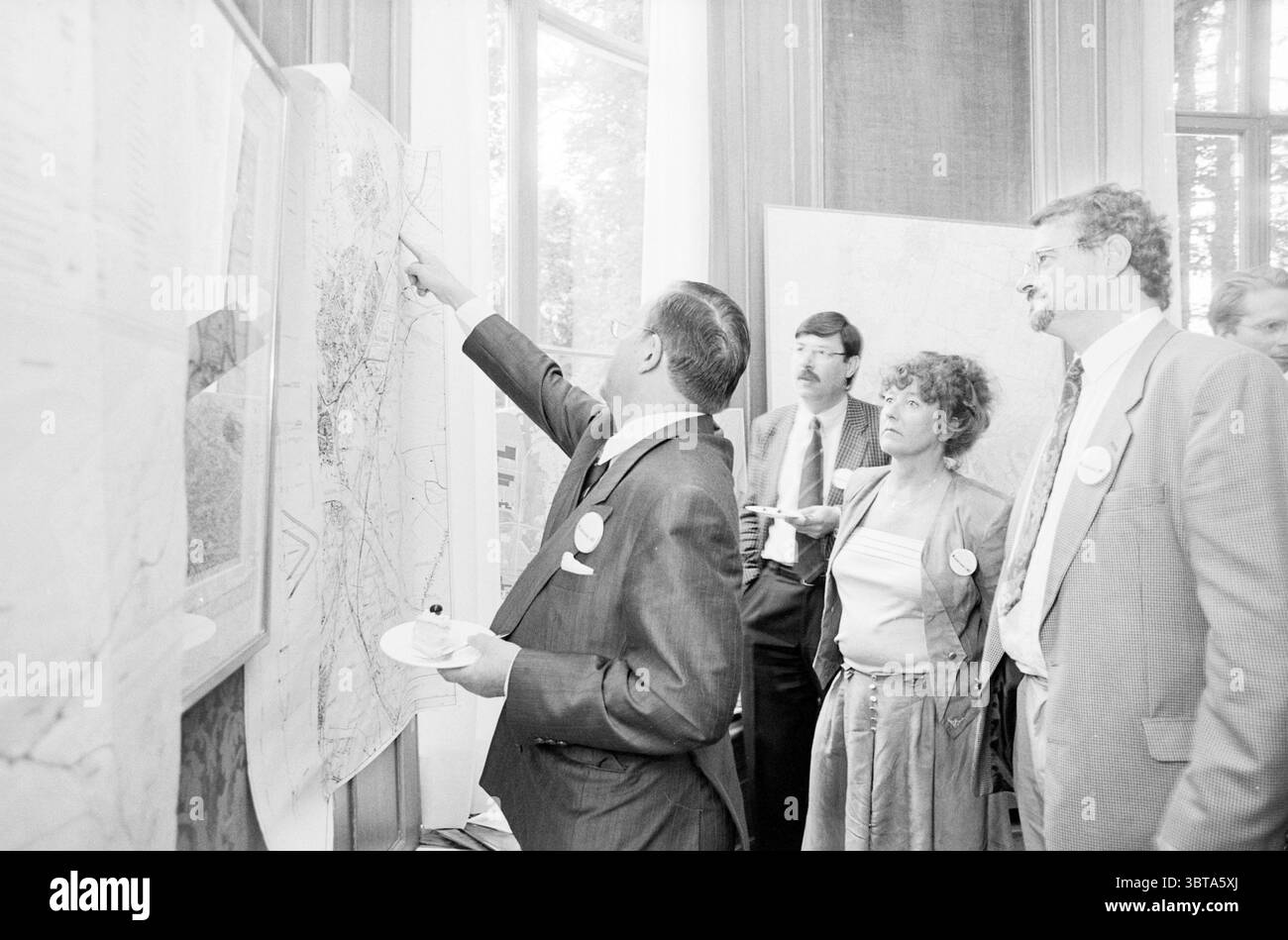 Meeting Wijkertunnel Beverwijk Beverwijk the Netherlands, Whizgle News, Dutch Desk, Niederlande, 1950 - 2000 am 02-07-1992. Das Bild enthält diese Themen. Die Szene zeigt eine Gruppe von Personen, die an einer Diskussion teilnehmen und in einem gut beleuchteten Raum stehen, der durch große Fenster gekennzeichnet ist, in denen natürliches Licht eindringen kann. Die Wände sind mit großen Karten und Diagrammen geschmückt, was auf eine detaillierte Planung oder Analyse hindeutet. Im Vordergrund bewegt sich ein Mann in einem scharfen, dunklen Anzug animiert auf eine der Karten, sein Ausdruck ist intensiv und konzentriert, während er Informationen oder Erkenntnisse mit Th teilt Stockfoto