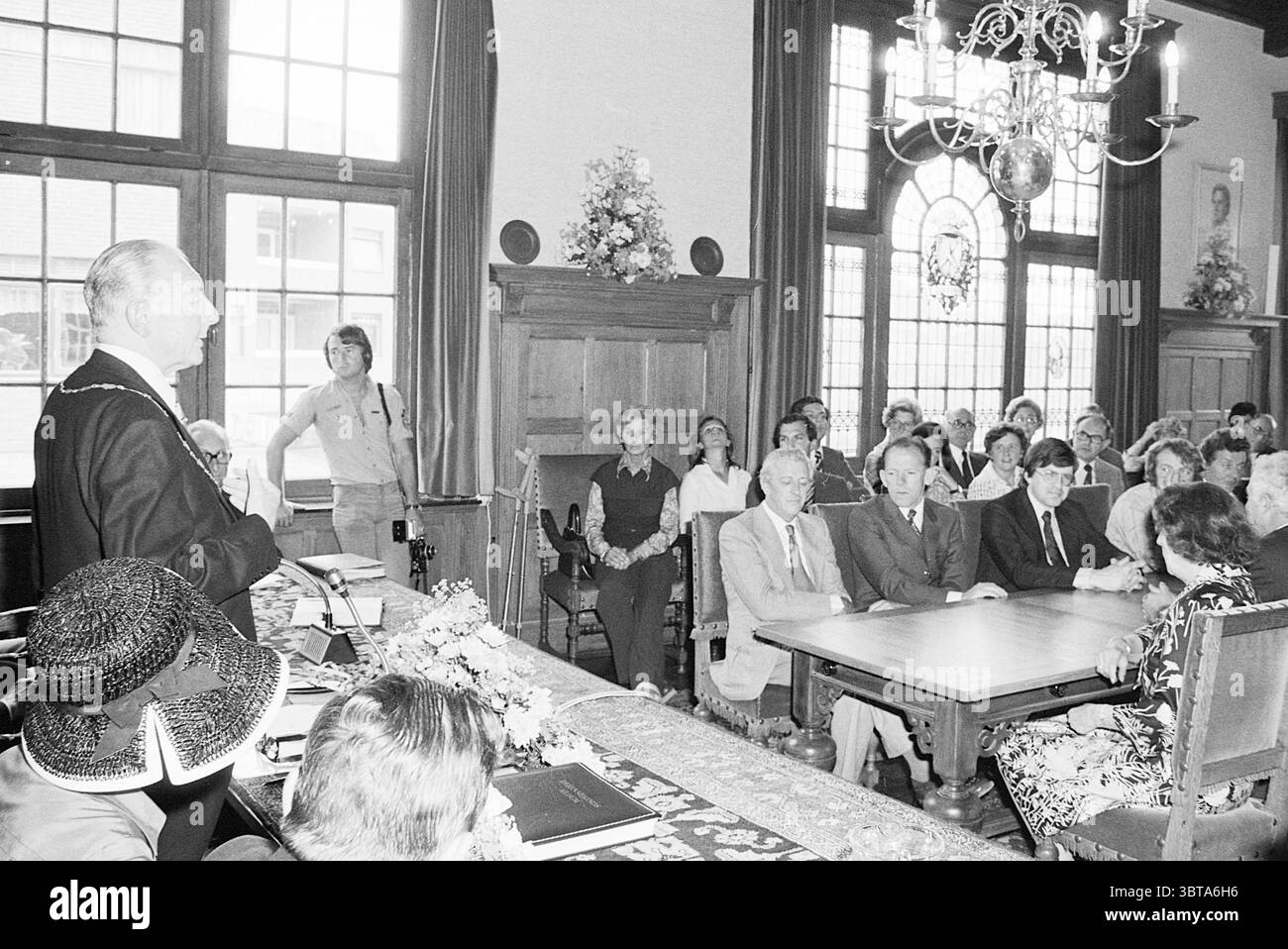 Abschied Bürgermeister Nawijn Juwel. Zandvoort Mayors, Whizgle News, Dutch Desk, Niederlande, 1950 - 2000 auf 30-06-1977. Das Bild enthält diese Themen. Die Szene entfaltet sich in einer formellen, eleganten Umgebung, die durch große Fenster gekennzeichnet ist, die natürliches Licht hereinlassen und den Teilnehmern ein warmes Leuchten verleihen. Das Zimmer hat ein klassisches Design mit kunstvollen Holzmöbeln, reichen Texturen und einem großen Tisch in der Mitte, der mit einem zarten Blumenarrangement verziert ist. An vorderster Front steht ein angesehener Mitarbeiter, der sich wahrscheinlich an das versammelte Publikum wendet. Seine formale Kleidung vermittelt Autorität und seine Stockfoto