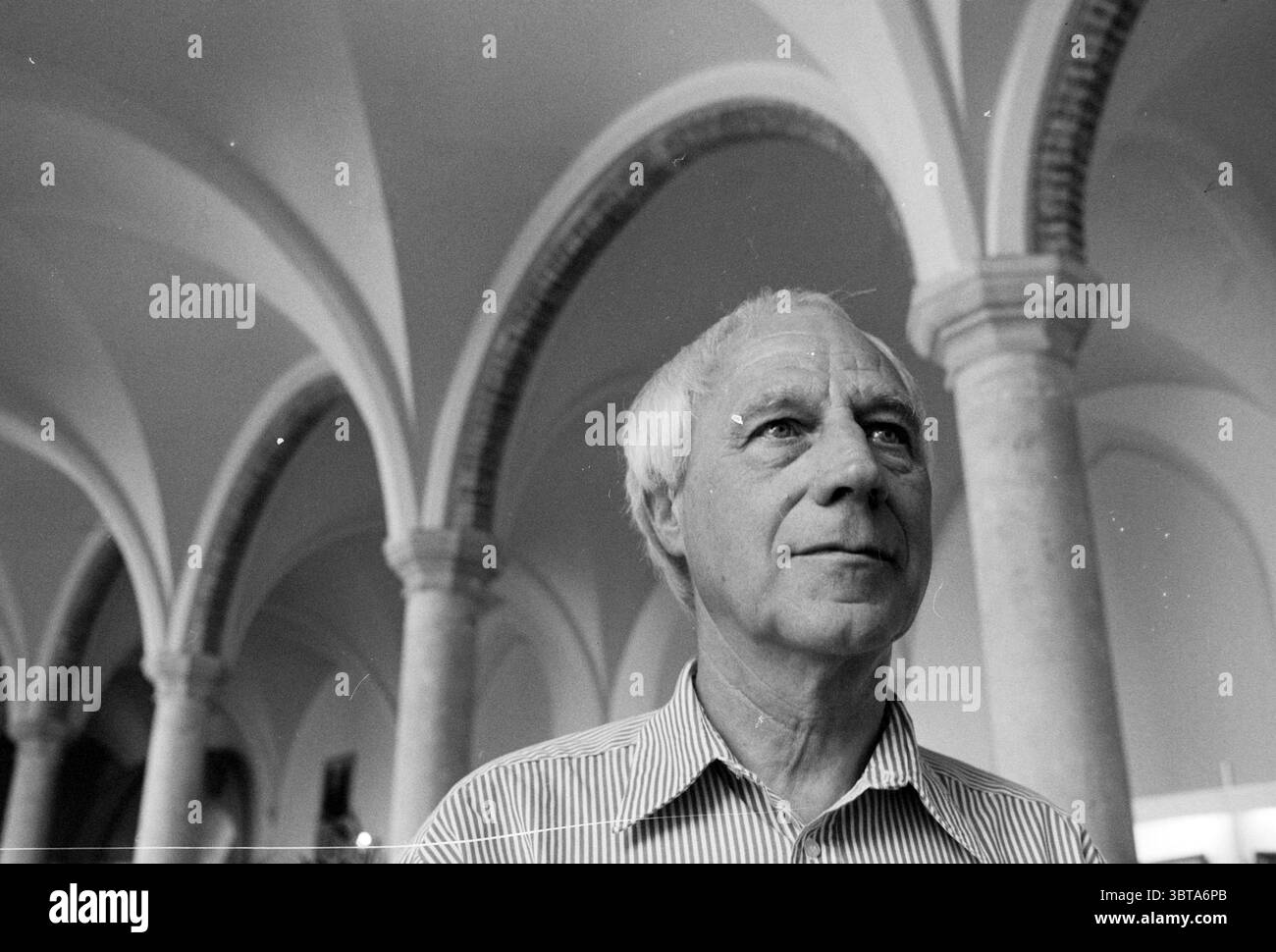 Künstler mr. Grabijn Rathaus Refektorium, Whizgle News, Dutch Desk, Niederlande, 1950 - 2000 am 03-08-1995. Diese Themen sind in der Abbildung dargestellt. In einer monochromatischen Szene steht ein Mann in einer weitläufigen, architektonischen Umgebung, die von hohen, anmutigen Bögen geprägt ist, die den Blick nach oben ziehen. Die Bögen aus Stein weisen ein klassisches Design auf und erzeugen einen Rhythmus, der der Umgebung ein Gefühl von Pracht verleiht. Der Ausdruck des Mannes ist nachdenklich, mit einer leichten Furche in der Stirn. Er trägt ein helles gestreiftes Hemd, das sich sanft mit den dunkleren Tönen des Hintergrunds abhebt. Sein Ha Stockfoto