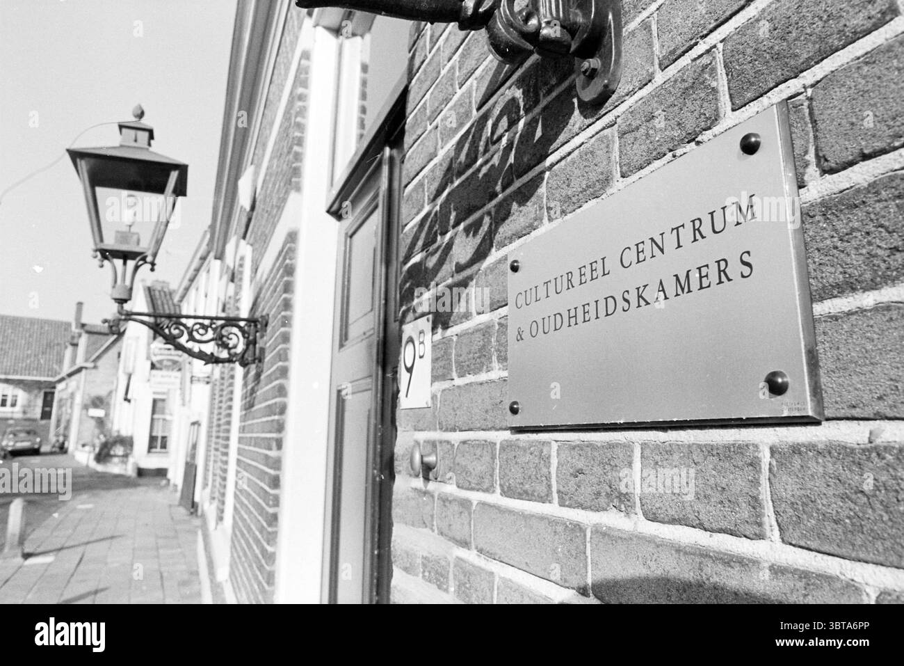 Eintritt zum Kulturzentrum Zandvoort Zandvoort Swaluëstraat, Whizgle News, Dutch Desk, Niederlande, 1950 - 2000 am 08-11-1978. Dies sind die Themen im Bild. In einer malerischen Straßenlandschaft dient eine Backsteinmauer als Kulisse, die mit einem markanten Schild in einem Winkel verziert ist. Die Oberfläche des Schildes weist eine polierte Textur auf, die sich im Kontrast zu den rauen, warmen Tönen der umliegenden Ziegelsteine, die in Braun- und Grautönen variieren, bildet. Links ist eine Vintage-Laterne an der Wand angebracht, deren dunkler Metallrahmen elegant nach außen geschwungen ist und ein weiches, atmosphärisches Leuchten in der Nähe erzeugt, was auf eine hindeutet Stockfoto