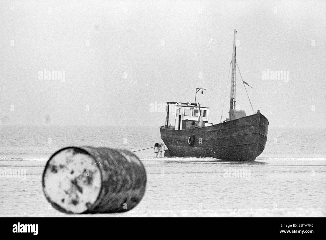 Stranded Ship Parnassia Beach Ships, Whizgle News, Dutch Desk, Niederlande, 1950 - 2000 am 16-10-1974. Dies sind die Elemente im Bild. Die Szene zeigt eine ruhige Küstenlandschaft, in der ein kleines Boot sanft am Ufer des Wassers liegt. Das Boot ist in dunklen Tönen lackiert und hat ein verwittertes Aussehen, was darauf hindeutet, dass es im Laufe der Zeit viele Gewässer durchquert hat. Seine Silhouette hebt sich vor einem trüben Hintergrund ab und fügt sich subtil in die gedämpften Farben der Szene ein. Im Vordergrund liegt ein großes Fass teilweise in den Sand getaucht. Der Zylinder ist durch die Zeit gekennzeichnet, seine Oberfläche weist Rostflecken und auf Stockfoto
