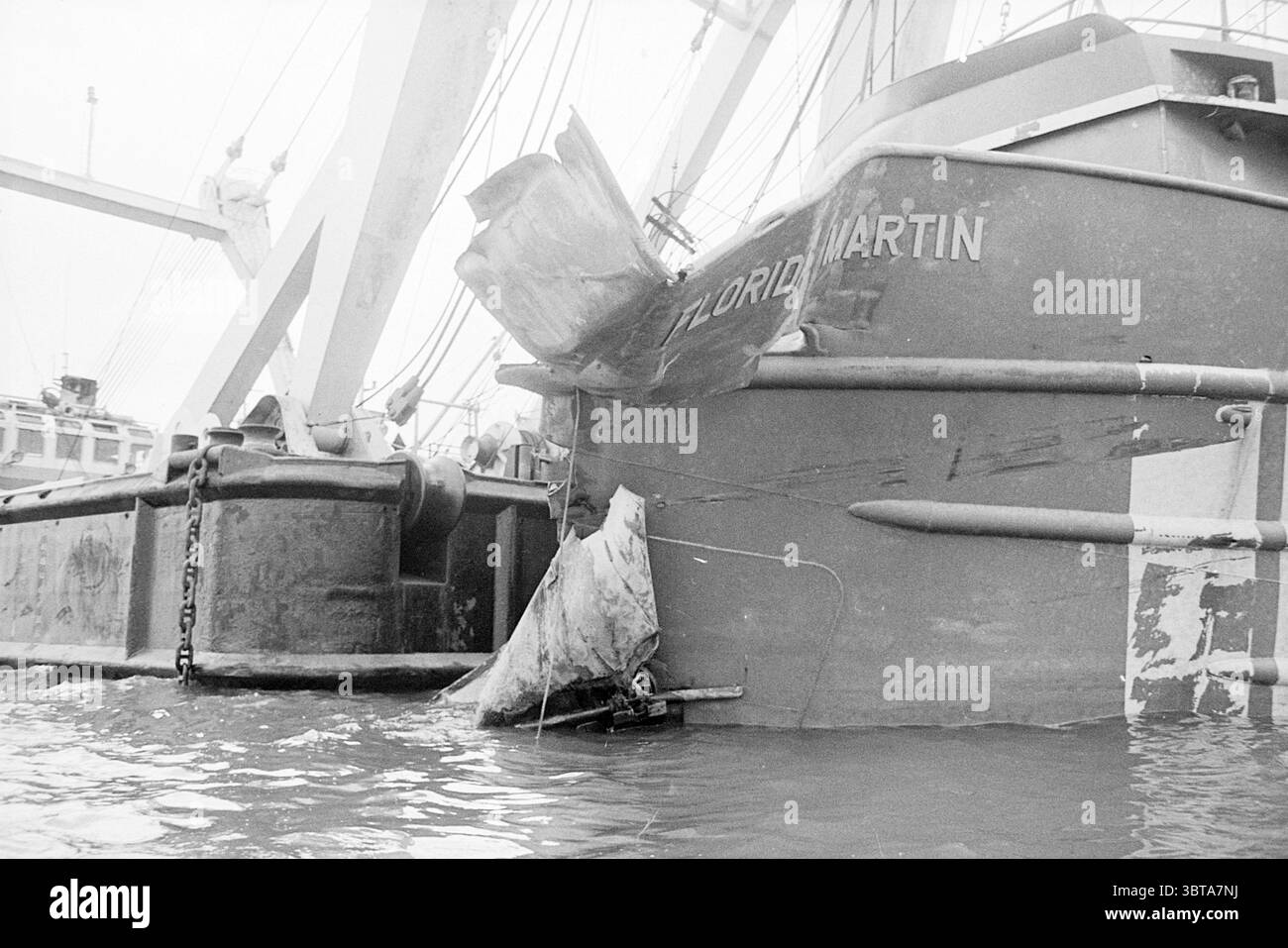 Bergungsschiff IJmuiden Ships, Whizgle News, Dutch Desk, Niederlande, 1950 - 2000 am 06-11-1974. Diese Themen sind in der Abbildung dargestellt. Die Szene zeigt ein großes Schiff, das an einem Hafen angedockt ist und eine robuste Schiffsstruktur zeigt, die sich durch sein industrielles Design auszeichnet. Der Rumpf des Schiffes ist überwiegend grau und hat Rostflecken, was darauf hindeutet, dass es viele Seefahrten überstanden hat. Der Bug ist deutlich beschädigt, wobei eine gezackte Kante an der Vorderseite sichtbar ist, was auf eine kürzliche Kollision oder Verschleiß hindeutet. Um das angedockte Schiff herum reflektiert das Wasser die gedämpften Farben des bewölkten Himmels und wirft ein wenig ein Stockfoto