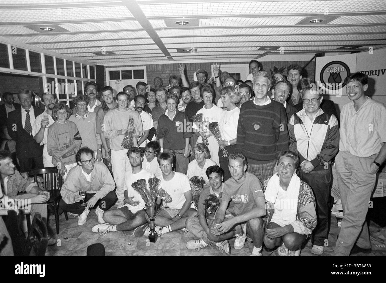 Preisträgerturnier, Whizgle News, Dutch Desk, Niederlande, 1950 - 2000 am 11.05.1990. Das Bild enthält diese Themen. In einem lebhaften Innenbereich versammelt sich eine große Gruppe von Menschen, die eine Atmosphäre des Feierns und der Kameradschaft ausstrahlen. Der Raum verfügt über eine strukturierte Decke mit hellen Lampen, die den Raum beleuchten und ein warmes und einladendes Leuchten erzeugen. Auf der einen Seite steht eine Mischung aus Männern und Frauen unterschiedlichen Alters zusammen, einige mit Trophäen und Blumensträußen, die auf eine jüngste Leistung oder Auszeichnung hinweisen. Die Teilnehmer tragen verschiedene Freizeitkleidung, darunter Sportbekleidung, h Stockfoto