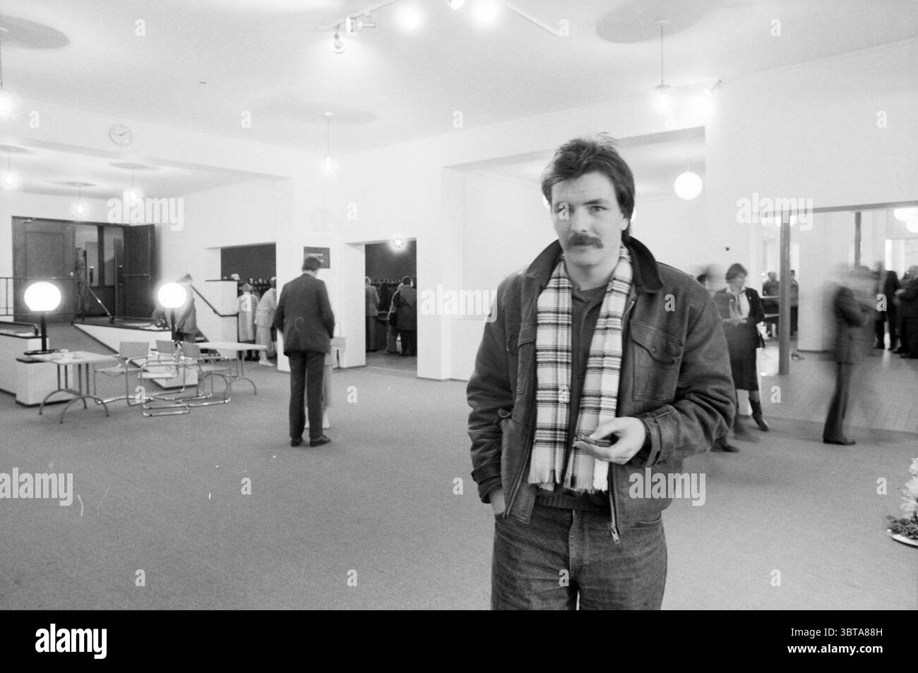 Int. Neue Konzerthalle Konzerthalle Konzerthalle Konzerthalle, Whizgle News, Dutch Desk, Niederlande, 1950 - 2000 am 21-02-1986. Dies sind die Themen im Bild. In einem geräumigen Innenbereich zeichnet sich die Szene durch ein raffiniertes, minimalistisches Design aus. Der Boden hat einen weitläufigen Teppich in einem neutralen Ton, der dezent mit den weißen Wänden kontrastiert. Weiche, diffuse Beleuchtung entsteht durch moderne, kugelförmige Leuchten, die von der Decke abhängen und eine warme und einladende Atmosphäre schaffen. Im Vordergrund steht ein Mann selbstbewusst, gekleidet in einer stilvollen dunklen Jacke mit hervorstehendem Kragen, gepaart mit Denim, an Stockfoto