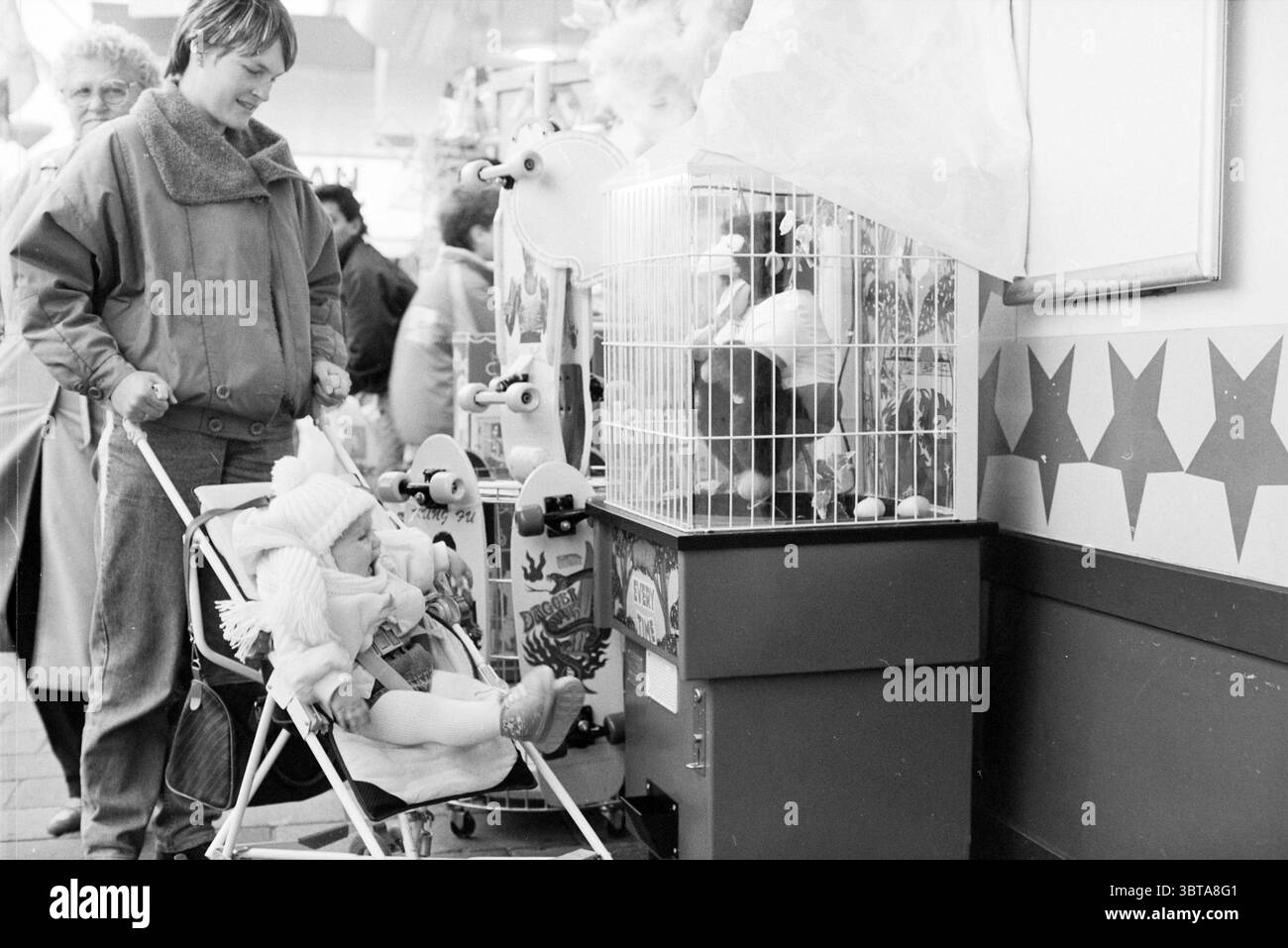 Toy Monkey bei Bart Smit IJmuiden Games and Toys IJmuiden the Netherlands, Whizgle News, Dutch Desk, Niederlande, 1950 - 2000 am 27-10-1988. Das Bild enthält diese Themen. Die Szene präsentiert einen belebten Innenraum, wahrscheinlich einen Marktplatz oder ein Messegelände, voller Menschen, die an verschiedenen Aktivitäten teilnehmen. Im Vordergrund zieht eine Frau in einer leichten Jacke in Begleitung eines Kindes im Kinderwagen die Aufmerksamkeit auf sich. Das Kind, warm gekleidet, mit einer Strickmütze und einem gemütlichen Outfit, scheint neugierig und konzentriert zu sein und blickt aufmerksam auf etwas in der Nähe. Daneben befindet sich ein kleiner Spielekabine, mit Stockfoto
