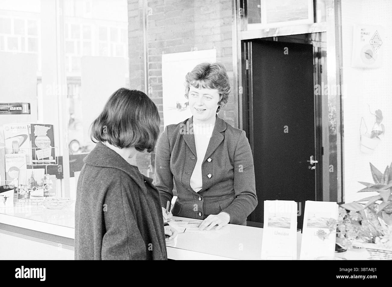 Frau Janssen V.O.O. IJmuiden Persons, Whizgle News, Dutch Desk, Niederlande, 1950 - 2000 am 19.12.1963. Das Bild enthält diese Themen. In einer hellen, luftigen Umgebung mit großen Fenstern, die viel Tageslicht einlassen, treffen zwei Frauen an der Rezeption miteinander. Der Schreibtisch ist elegant und modern, mit einfachen Broschüren und Informationen über verschiedene Dienstleistungen. Die Frau hinter dem Schreibtisch hat ein freundliches Auftreten und ein warmes Lächeln, das die einladende Atmosphäre noch verstärkt. Sie hat ein kurzes, leicht lockiges Haar und trägt einen dunklen Blazer über einem hellen Oberteil, was ihr eine gute Figur macht Stockfoto