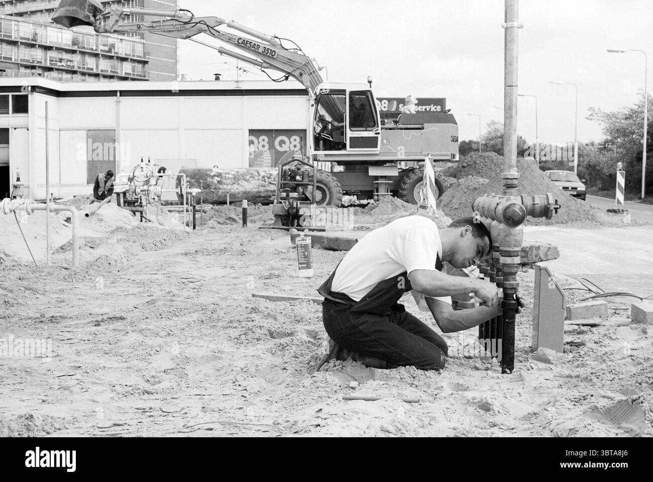 Aktivitäten Q8 IJmuiden IJmuiden the Netherlands, Whizgle News, Dutch Desk, Niederlande, 1950 - 2000 am 08-09-1992. Die Abbildung zeigt diese Themen. Die Szene fängt eine Baustelle voller Aktivität ein. Im Vordergrund kniet ein Arbeiter auf sandigem Boden und konzentriert sich intensiv auf eine Rohrverschraubung. Er trägt ein dunkles Outfit, mit einem deutlichen Kontrast zum hellen Sand unter ihm. Seine Haltung deutet auf Konzentration und Fleiß hin, während er die Pfeife justiert. Hinter ihm sind große Maschinen stationiert, darunter ein prominenter Bagger, der den Hintergrund dominiert. Sein metallischer Körper glänzt unter Stockfoto