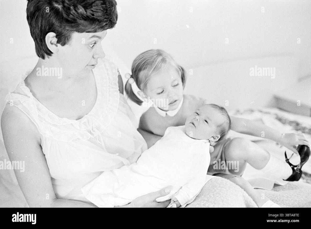 Mutter mit Kleinkind und Baby. Whizgle News, Dutch Desk, Niederlande, 1950 - 2000. Diese Themen werden in der Abbildung angezeigt. In einem sanft beleuchteten Innenraum sitzt eine Frau auf einem hellen Bett. Ihr Ausdruck ist eine Mischung aus Überraschung und Zuneigung, während sie nach rechts blickt. Ihr kurzes, dunkles Haar umrahmt ihr Gesicht, das mit sanftem, diffusem Licht beleuchtet wird, was ihre Gesichtszüge und die Emotionen unterstreicht. Sie trägt ein weiches, weißes Kleidungsstück mit zarten Rüschen, die ihrem Aussehen ein Gefühl von Weichheit verleihen. In ihren Armen ist ein Kleinkind, das in eine helle Decke gehüllt ist, ihr winziges Gesicht Stockfoto