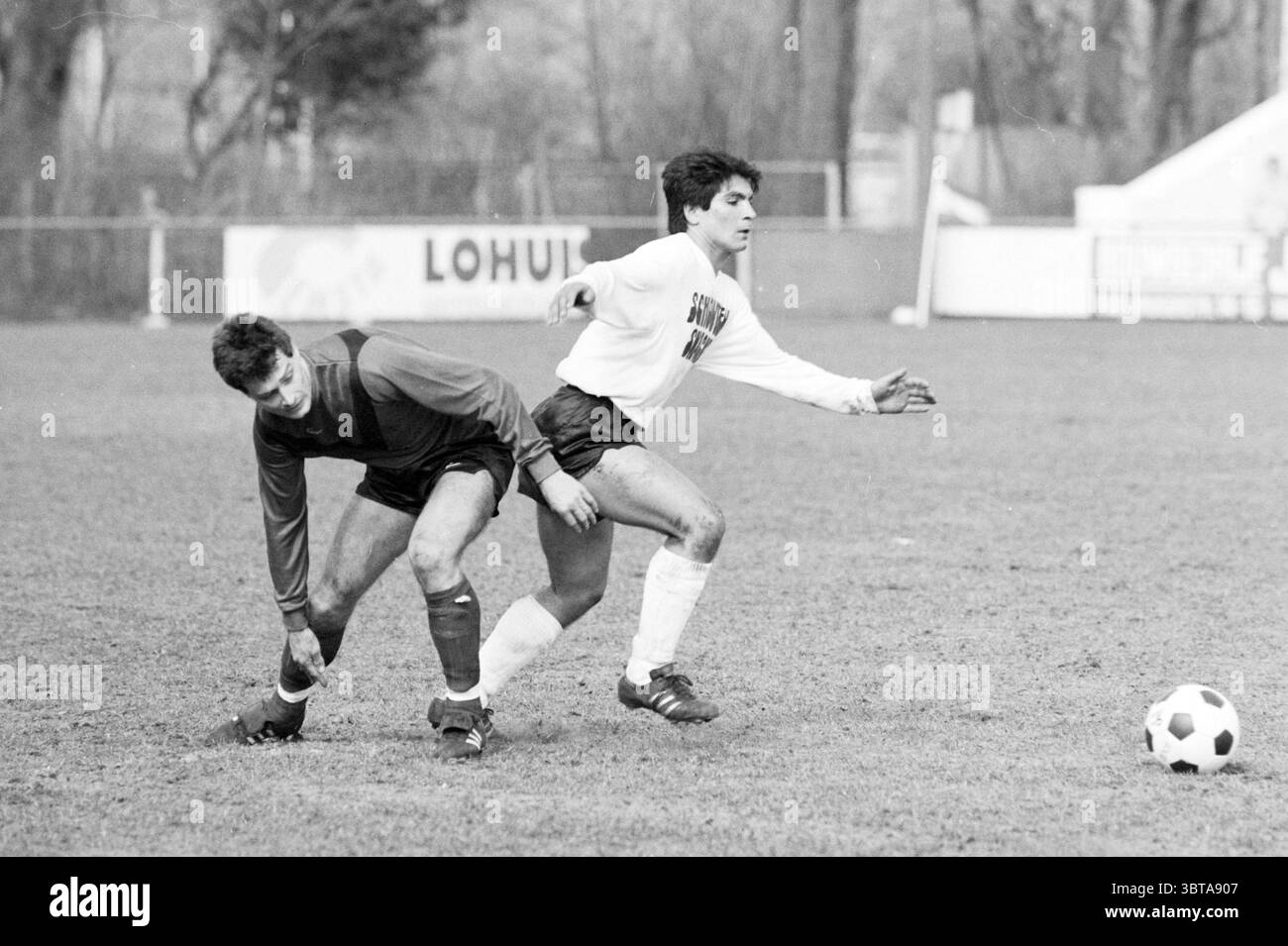 SMS - Aalsmeer Football, Whizgle News, Dutch Desk, Niederlande, 1950 - 2000 am 29-03-1987. Die Abbildung zeigt diese Themen. Die Szene fängt einen dynamischen Moment auf einem grasbewachsenen Fußballfeld ein. Zwei Athleten stehen im Mittelpunkt der Action, die sich um den Besitz des Balls kämpfen. Auf der linken Seite lehnt sich ein Spieler, der in einem dunklen Trikot mit Shorts gekleidet ist, leicht in der Hocke nach vorne, als er versucht, sein Gleichgewicht wiederherzustellen. Sein Ausdruck zeigt Entschlossenheit, und er scheint sich auf den Ball vor ihm zu konzentrieren. Der zweite Spieler, der ein helles Langarmshirt und Shorts trägt, ist in der Mitte Stockfoto