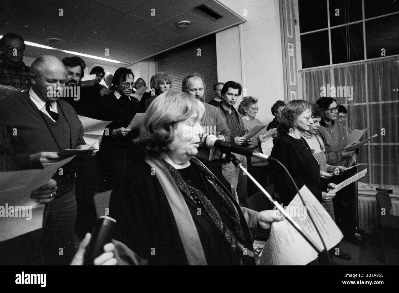 Singen Sie Protest von Nachbarschaftsräten im Rathaus mit Bürgermeister Schmitz Haarlem the Netherlands, Whizgle News, Dutch Desk, Niederlande, 1950 - 2000 am 24.11.1991. Dies sind die Elemente im Bild. Die Szene fängt eine Gruppe von Individuen ein, die sich in einem etwas schwach beleuchteten Raum versammelt haben, mit einem Fokus auf eine Frau, die an der Vorderseite steht. Sie befindet sich etwas rechts von der Mitte, hält ein Mikrofon und blickt auf das Publikum hin und strahlt ein Gefühl von Autorität und Präsenz aus. Die Frau scheint die Gruppe zu führen, die aus Männern und Frauen unterschiedlichen Alters besteht, von denen viele halten Stockfoto