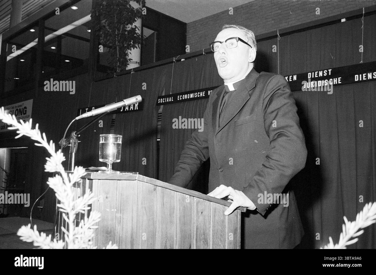 Jahrestagung 60 Jahre LTB Treslong Hillegom LTB Meetings, Whizgle News, Dutch Desk, Niederlande, 1950 - 2000 am 17-12-1975. Diese Themen werden in der Abbildung angezeigt. In einem Raum mit dunkler Kulisse steht ein Mann auf einem hölzernen Podium und spricht leidenschaftlich. Seine Kleidung besteht aus einem maßgeschneiderten schwarzen Anzug, der den ernsten Ton der Szene unterstreicht. Eine dicke, schwarze Brille liegt auf seiner Nase, die einen konzentrierten und intensiven Ausdruck bildet, der das Publikum wahrscheinlich fesseln wird. Zu seiner Linken fängt ein Glas mit einer klaren Flüssigkeit das Licht auf, was auf ein Getränk hindeutet, das erfrischend sein könnte. Stockfoto