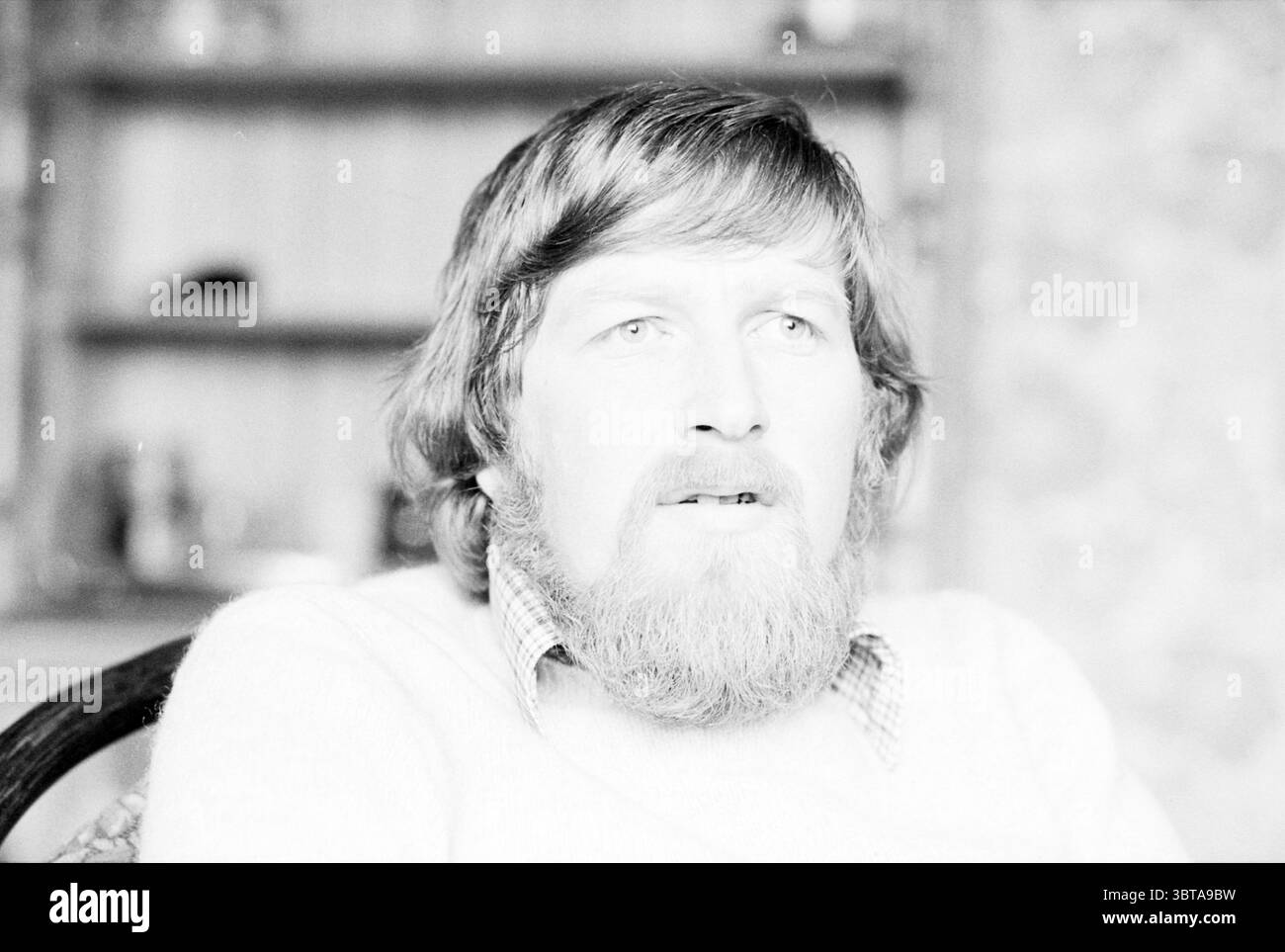Porträt von Mr. Mak, der mit alten Flugzeugen reist, Whizgle News, Dutch Desk, Niederlande, 1950 - 2000 am 05-02-1980. Die Abbildung zeigt diese Themen. In der Szene steht ein Mann mit einem vollen, gut gepflegten Bart und mittellangen Haaren im Mittelpunkt. Sein Ausdruck vermittelt ein Gefühl der Besinnung oder Überraschung, mit breiten Augen, die Aufmerksamkeit auf sich ziehen. Er trägt einen hellen Pullover über einem karierten Hemd, der seinem Outfit eine dezente Textur verleiht. Der Hintergrund weist weiche, unscharfe Elemente auf, die auf eine Wohnumgebung, möglicherweise ein Wohnzimmer oder ein Arbeitszimmer hinweisen. Es gibt Hinweise auf Regale Stockfoto