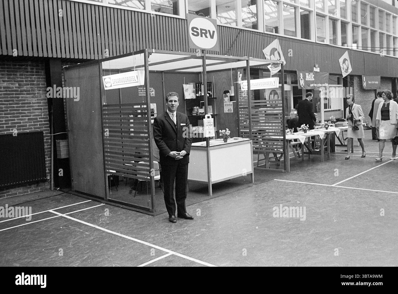 Molkereiausstellung Badminton Hall Exhibitions, Whizgle News, Dutch Desk, Niederlande, 1950 - 2000 am 01-05-1968. Diese Themen sind in der Abbildung dargestellt. Die Szene präsentiert eine geschäftige Innenumgebung mit einer Vielzahl von Kabinen, die in einem hallenartigen Raum angeordnet sind. Die Wände verfügen über große Fenster, durch die natürliches Licht den Bereich durchflutet und eine helle und einladende Atmosphäre geschaffen wird. Der Boden hat eine polierte, möglicherweise hölzerne Oberfläche, und es gibt markierte Linien, die auf einen Sportplatz hinweisen, was auf eine multifunktionale Nutzung des Ortes hindeutet. Im Mittelpunkt der Szene steht ein Holzstand mit einem stromlinienförmigen Design, fla Stockfoto