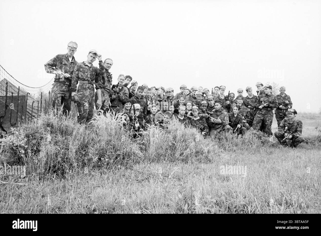 Paintball Shooting Sloterdijk, Whizgle News, Dutch Desk, Niederlande, 1950 - 2000 am 06-06-1992. Dies sind die Elemente im Bild. Die Szene zeigt eine große Gruppe von Personen, insgesamt etwa dreißig, die zusammen in einer Umgebung im Freien stehen. Sie tragen überwiegend Tarnkleidung im Militärstil, die in Grün-, Braun- und Schwarztönen variiert. Die Kleidung deutet auf eine Verbindung zu einem Militär oder einer Trainingsumgebung hin. Die Gruppe ist in einer halblässigen Formation angeordnet, wobei einige Individuen stehen und andere auf einer grasbewachsenen Fläche knien oder hocken, die etwas erscheint Stockfoto