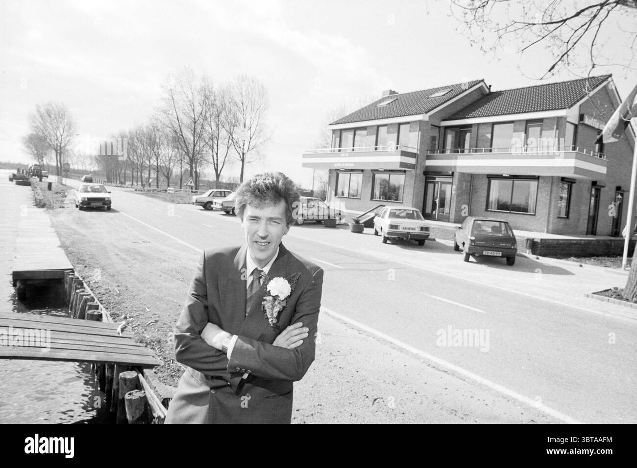 Mr. Short für Café Leimuiderbrug Café Personen, Whizgle News, Dutch Desk, Niederlande, 1950 - 2000 am 15-03-1989. Dies sind die Themen im Bild. Die Szene präsentiert eine Straßenlage, die durch eine Mischung aus urbanen und pastoralen Elementen gekennzeichnet ist. Ein gut gepflegtes zweistöckiges Gebäude mit einem gekachelten Dach und großen Fenstern, die die Umgebung reflektieren, steht gut sichtbar. Das Gebäude erinnert an eine Wohn- oder Geschäftsnutzung und verbindet moderne Architektur mit funktionalem Design. Im Vordergrund steht ein Gentleman selbstbewusst mit überkreuzten Armen. Er trägt einen formellen Anzug, der mit einem li verziert ist Stockfoto