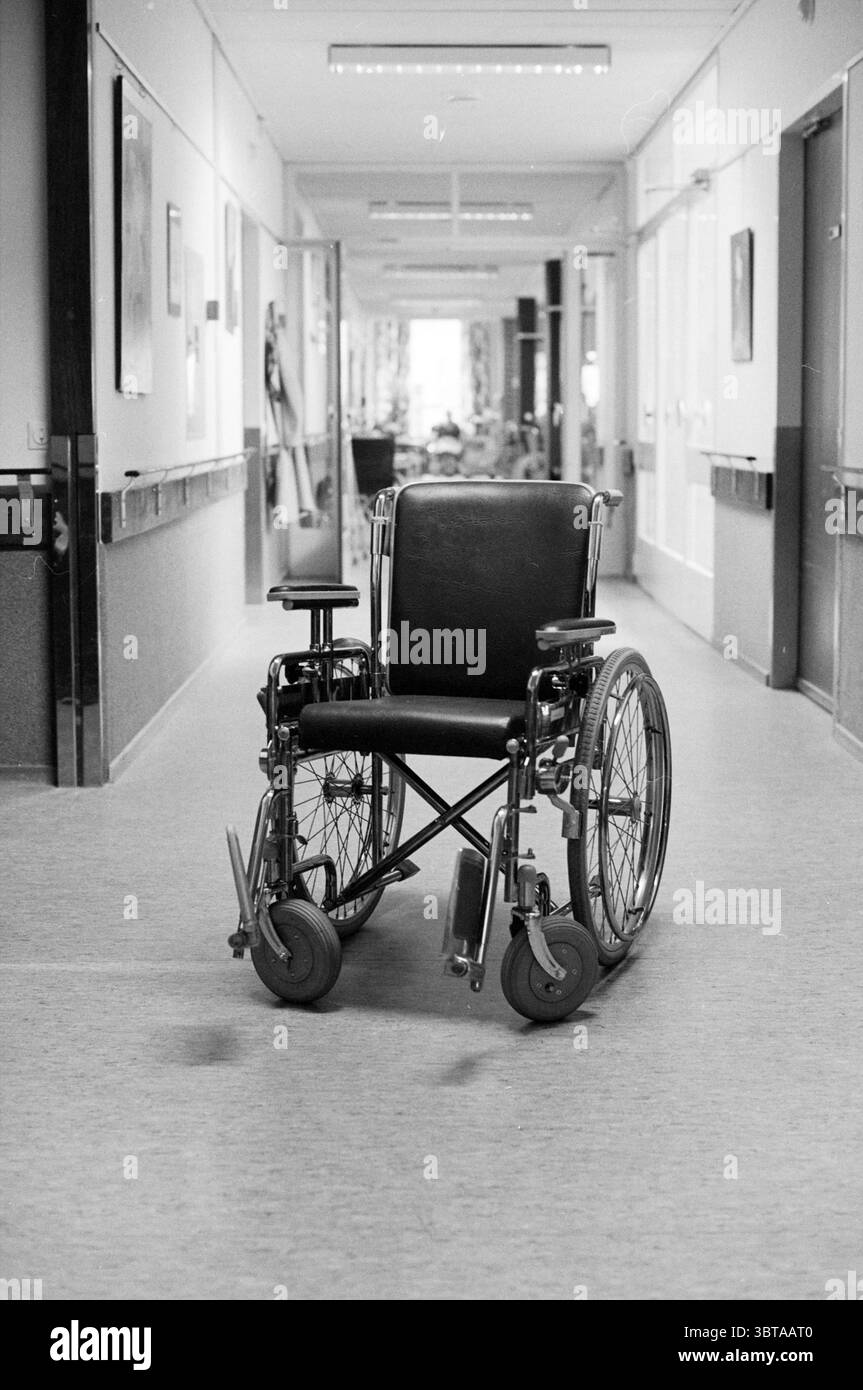 Zuiderhout ein Rollstuhl in einem Flur Haarlem the Netherlands, Whizgle News, Dutch Desk, Niederlande, 1950 - 2000 am 12-10-1988. Diese Themen werden in der Abbildung angezeigt. Die Szene zeigt einen langen, schmalen Korridor innerhalb einer Gesundheitseinrichtung, der sich durch sein institutionelles Design auszeichnet. Der Boden ist gedämpft, hell gefärbt und trägt zu einer sauberen, aber sterilen Atmosphäre bei. Auf beiden Seiten des Korridors kann man teilweise sichtbare Räume oder Bereiche sehen, die auf die Aktivität im Inneren hinweisen, obwohl die Details unscharf bleiben. Im Zentrum der Komposition steht ein Einzelrollstuhl, sein metallischer Stockfoto