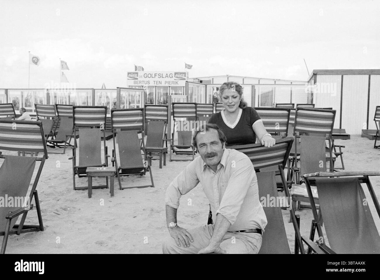 Strandmieter Bertus Ten Pierik Zandvoort Strand und Strände, Whizgle News, Dutch Desk, Niederlande, 1950 - 2000 auf 08-08-1978. Das Bild enthält diese Themen. Die Szene fängt eine Strandkulisse ein, wo eine helle, sandige Landschaft mit Reihen gestreifter Liegestühle gefüllt ist. Die Stühle in wechselnden Farben von dunklem und hellem Holz erzeugen ein rhythmisches Muster im Vordergrund. In der Mitte sitzt ein Mann mit einer entspannten Haltung. Er hat ein helles Hemd und einen unverwechselbaren Schnurrbart, der eine lässige Atmosphäre verkörpert. Sein Ausdruck deutet auf ein Gefühl des Genusses oder Lachens hin. Hinter ihm steht eine Frau Stockfoto