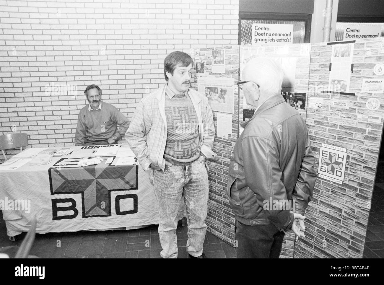 Erste-Hilfe-Demonstration Skagerhof Einkaufszentrum Hoofddorp erste Hilfe, Whizgle News, Dutch Desk, Niederlande, 1950 - 2000 auf 30-10-1987. Die Abbildung zeigt diese Themen. Die Szene fängt eine geschäftige Innenumgebung mit einer Atmosphäre des gemeinschaftlichen Engagements ein. Im Vordergrund stehen zwei Männer, die sich unterhalten. Einer, der lässig in einem gemusterten Pullover und einer Jeans gekleidet ist, zeigt ein freundliches Auftreten, während er sich leicht nach vorne lehnt. Der andere Mann, ein älteres Individuum, trägt eine dunkle Jacke und Brille und hört aufmerksam zu. Hinter ihnen ist ein Tisch mit einem Tuch bedeckt und präsentiert eine Sammlung von Stockfoto