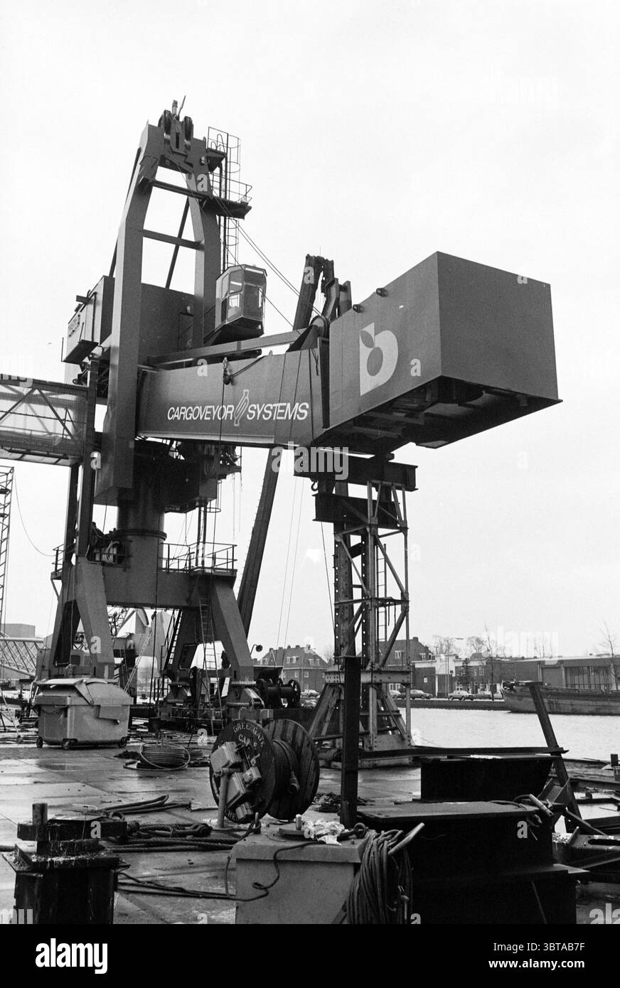 Neuer Kran Figee Figee Cranes Haarlem Niederlande, Whizgle News, Dutch Desk, Niederlande, 1950 - 2000 am 05-02-1980. Diese Themen werden in der Abbildung angezeigt. Die Szene zeigt einen großen Industriekran in der Nähe eines Wasserkörpers, vermutlich Teil eines Ladungsbereichs. Der Kran ist robust und metallisch, mit einer Mischung aus dunkelgrauen und schwarzen Tönen, die seine Struktur dominieren. Die Maschine hat eine etwas eckige und funktionale Bauweise, die auf ihre hohe Beanspruchung in einer industriellen Umgebung hinweist. Im Vordergrund befinden sich verschiedene Elemente wie Seile und Kabel, die lose herabhängen und auf ein hindeuten Stockfoto