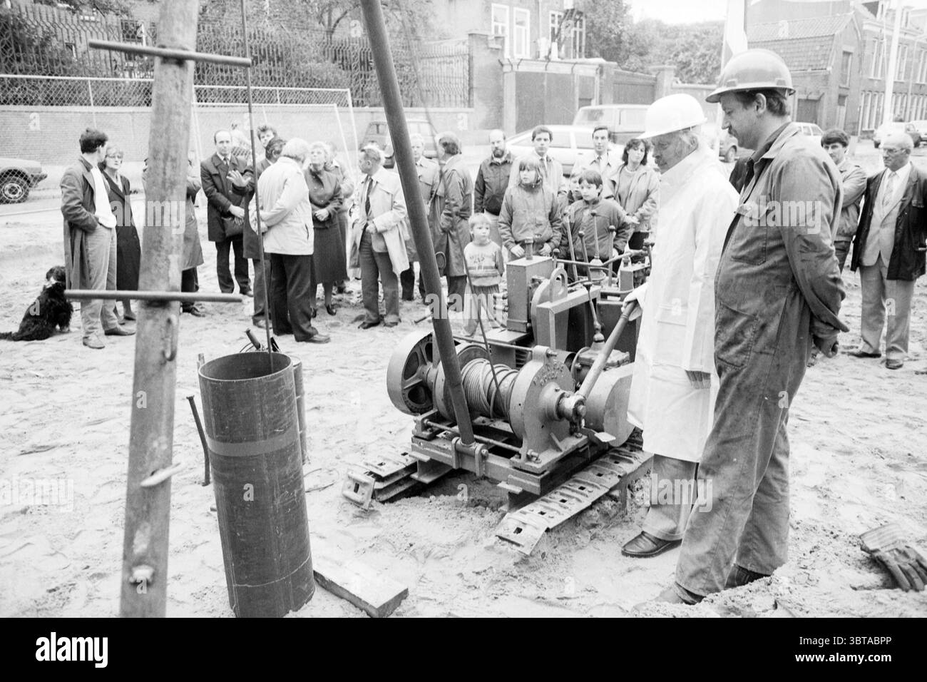 Beginn der 2. Bauphase Spaarnwouderstraat Start Haarlem Spaarnwouderstraat Niederlande, Whizgle News, Dutch Desk, Niederlande, 1950 - 2000 am 17-10-1985. Diese Themen werden in der Abbildung angezeigt. Die Szene zeigt eine Baustelle mit einer vielfältigen Gruppe von Menschen, die sich um eine Maschine versammelt haben. Im Vordergrund befinden sich zwei Arbeiter neben einem großen, industriell wirkenden Gerät, möglicherweise einem Bohrgerät. Ein Arbeiter trägt weiße Schutzkleidung, einschließlich Helm, während der andere einen dunklen Mantel trägt und einen Helm trägt. Beide haben aufmerksame Ausdrücke, Stockfoto