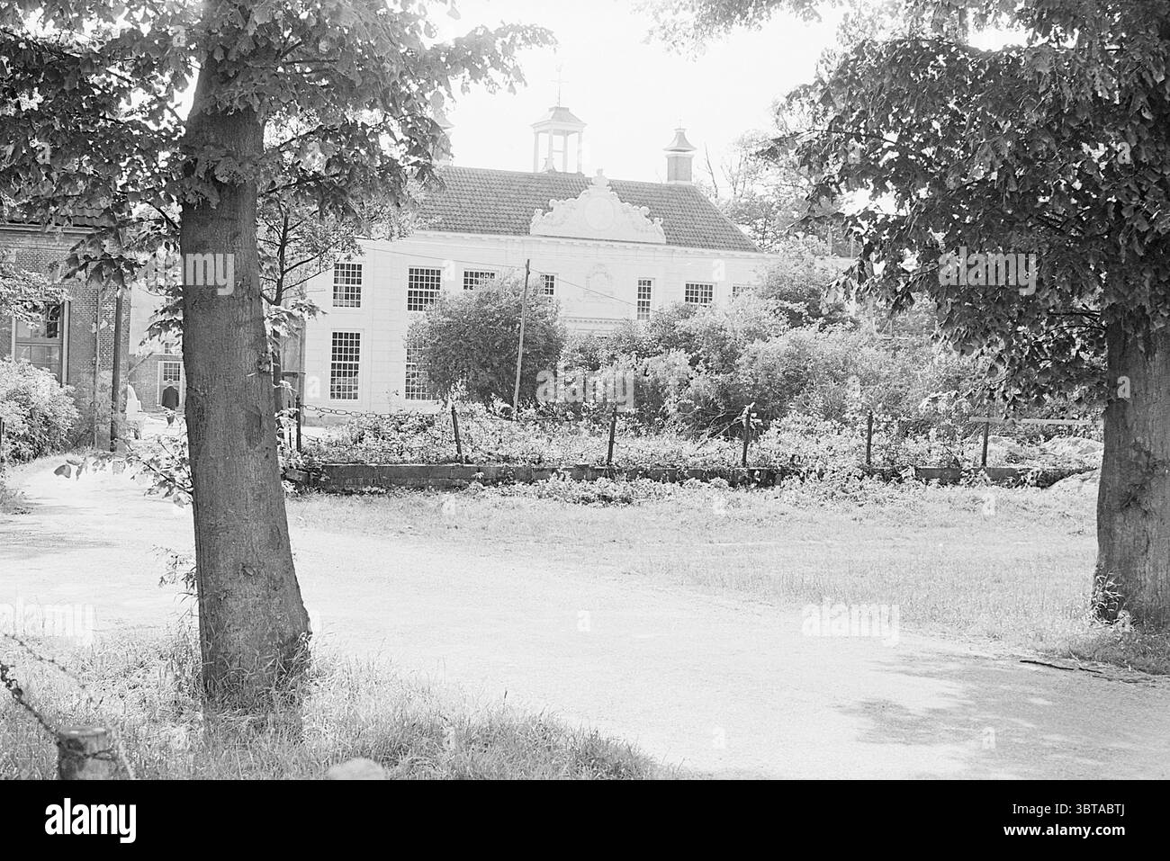 Ruin Bekenstein restaurierte Gebäude Restauration Ruin Velsen-Zuid Rijksweg A9, Whizgle News, Dutch Desk, Niederlande, 1950 - 2000 am 16-06-1964. Diese Themen werden in der Abbildung angezeigt. Die Szene bietet eine ruhige Umgebung im Freien, dominiert von einem gut gepflegten Gebäude, das sich durch eine weiße Fassade und eine spitzen Dachkonstruktion mit markanten Kuppeln an den Kanten auszeichnet. Die Struktur ist umgeben von üppigen grünen Pflanzen und dichten Laubbäumen, die zu einer lebhaften und dennoch ruhigen Atmosphäre beitragen. Im Vordergrund bilden zwei große Bäume mit strukturierten, belaubten Vordächern die Szene, deren Stämme robust und en sind Stockfoto